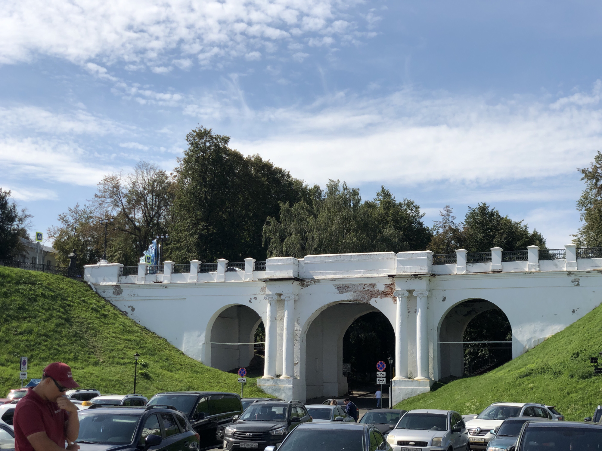 A white arched bridge spans across a grassy slope under a partly cloudy blue sky. The bridge has three visible arches, with the central arch appearing larger. The bridge's facade shows signs of wear, with some brickwork exposed. A black metal railing tops the bridge. Green grass covers the sloped embankment on either side of the bridge. Trees with dark green foliage are visible behind the bridge. In the foreground, several cars are parked. A man wearing a red cap and shirt stands near the left side of the frame, looking down. The scene appears to be located at the Strelka (Arrow) spit in Yaroslavl, Russia, a prominent historical and architectural landmark where the Volga and Kotorosl rivers meet.