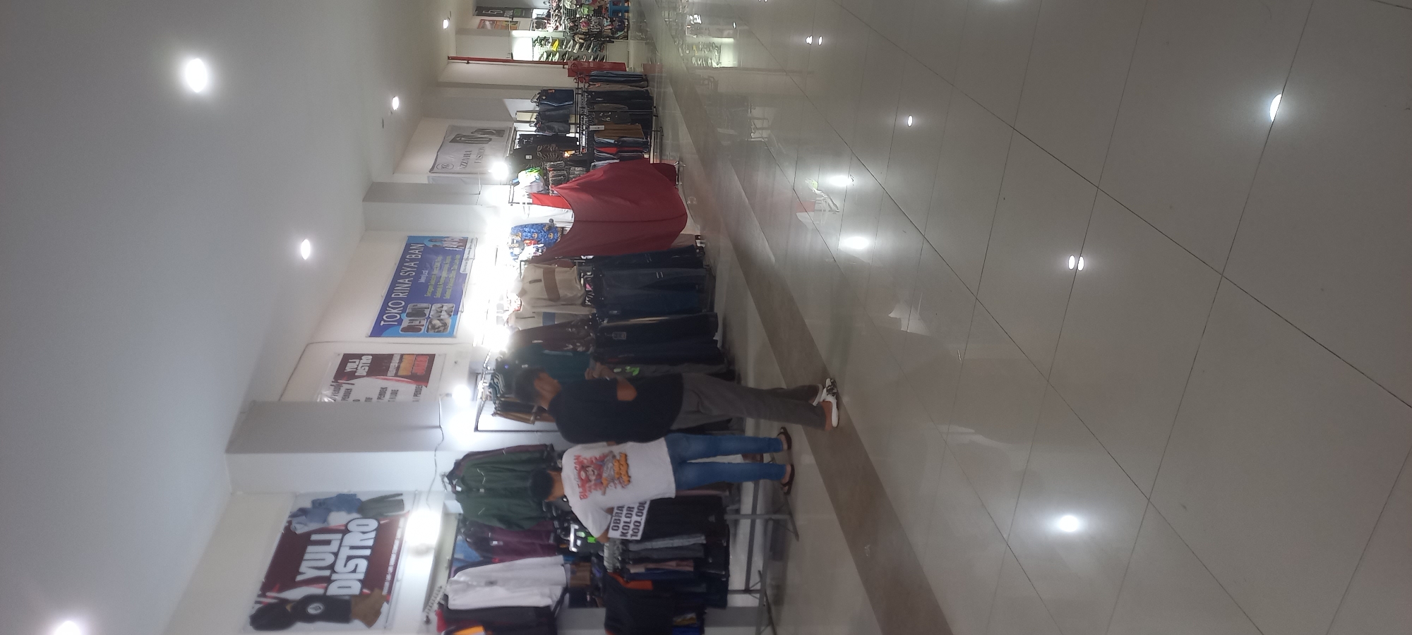 The image depicts the interior of a clothing store, likely in Melayu, Indonesia. Two individuals, both male, are visible. One, wearing a white t-shirt with an image and text, and blue jeans, stands with his back to the camera, looking at clothes displayed on a rack. The other, in a black t-shirt and grey pants, is also looking at clothes. The store features numerous clothing racks filled with various garments, including jackets, shirts, and pants. Several signs are visible on the walls, including "YULI DISTRO" and "TOKO RINA SYA'BAN." The floor is tiled and highly reflective, showing overhead lighting. The overall environment suggests a commercial space selling apparel.