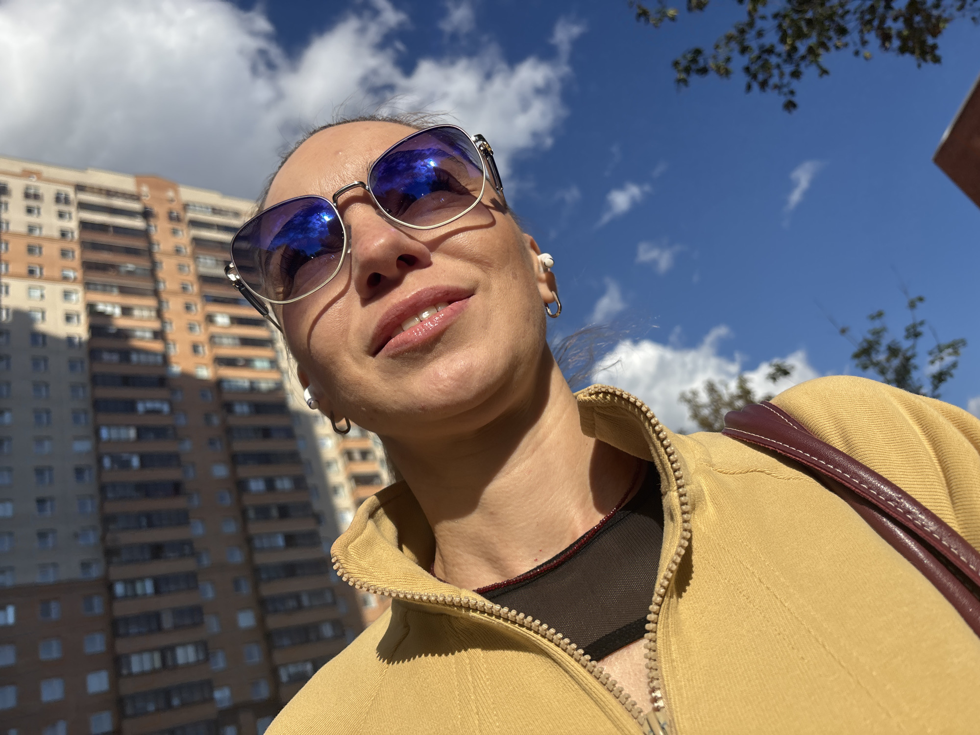 A woman is taking a selfie outdoors, with a tall brick building in the background and a bright blue sky with fluffy white clouds above. She is wearing a yellow zippered jacket, sunglasses with blue lenses, and a black top with a visible strap detail. The sun is casting a strong shadow on her face. The image is oriented vertically, giving the impression that the camera is tilted upwards.