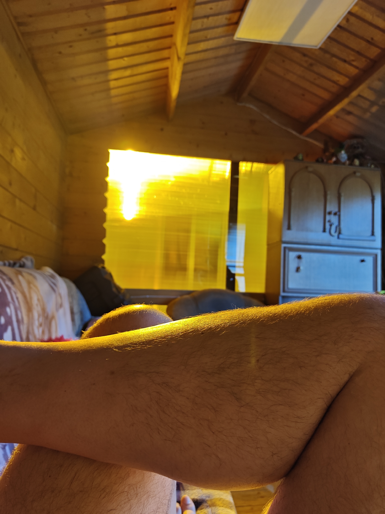 The image depicts a close-up view of a person's legs in a dimly lit wooden room. The legs are illuminated by warm, golden sunlight streaming through a window covered with yellow material. The sunlight highlights the texture of the skin and the presence of body hair. In the background, a bed with a patterned duvet is visible, along with a wooden cabinet. The ceiling is made of wooden beams. The location is not specified within the image or its context.