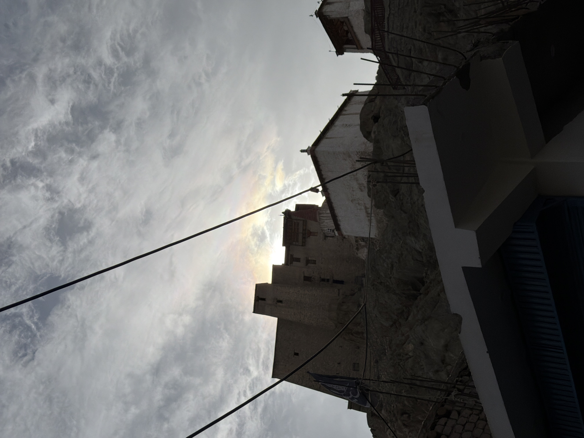 The image shows a view of the Leh Palace, a 17th-century royal palace in Leh, Ladakh, India. The palace is a towering structure built on a hilltop, with its stone walls and multiple stories visible against a cloudy sky. A faint rainbow or atmospheric optical phenomenon is visible in the sky above the palace. In the foreground, there are wires strung across the frame, and parts of a building under construction or renovation are visible. The overall scene depicts a historical landmark in a mountainous region.