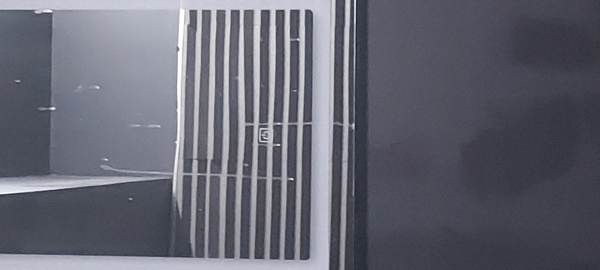 The image shows a corner of a public restroom, likely in Tashkent, Uzbekistan. A dark gray partition is visible on the right. A stall door, made of vertical black and white bars with a small square enclosure with the letter "F" on it, is on the left. Another dark gray partition is visible behind the door. There appears to be a sink basin also.
