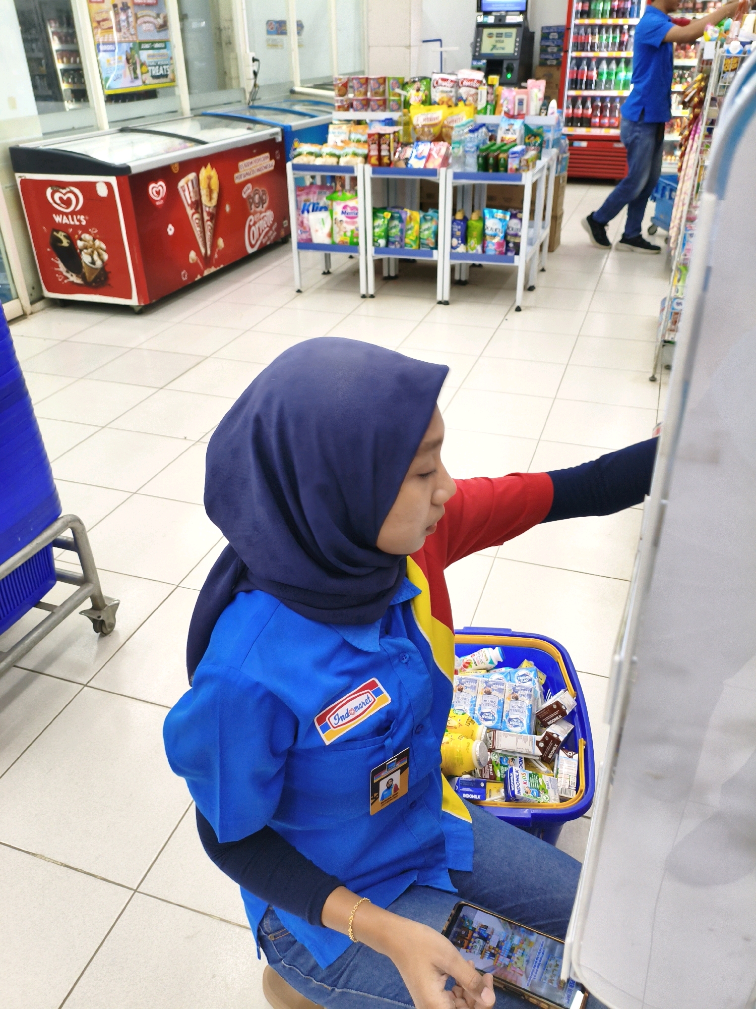 A young woman wearing a blue uniform with a red and yellow stripe, and a dark blue hijab, is in a convenience store in Balesono, Indonesia. She is kneeling, holding a smartphone in her right hand and a blue shopping basket filled with various packaged goods in her lap. The basket contains cartons of milk, snacks, and other grocery items. In the background, shelves are stocked with products, and a man in a blue shirt and jeans is standing near a refrigerator. The floor is tiled white.
