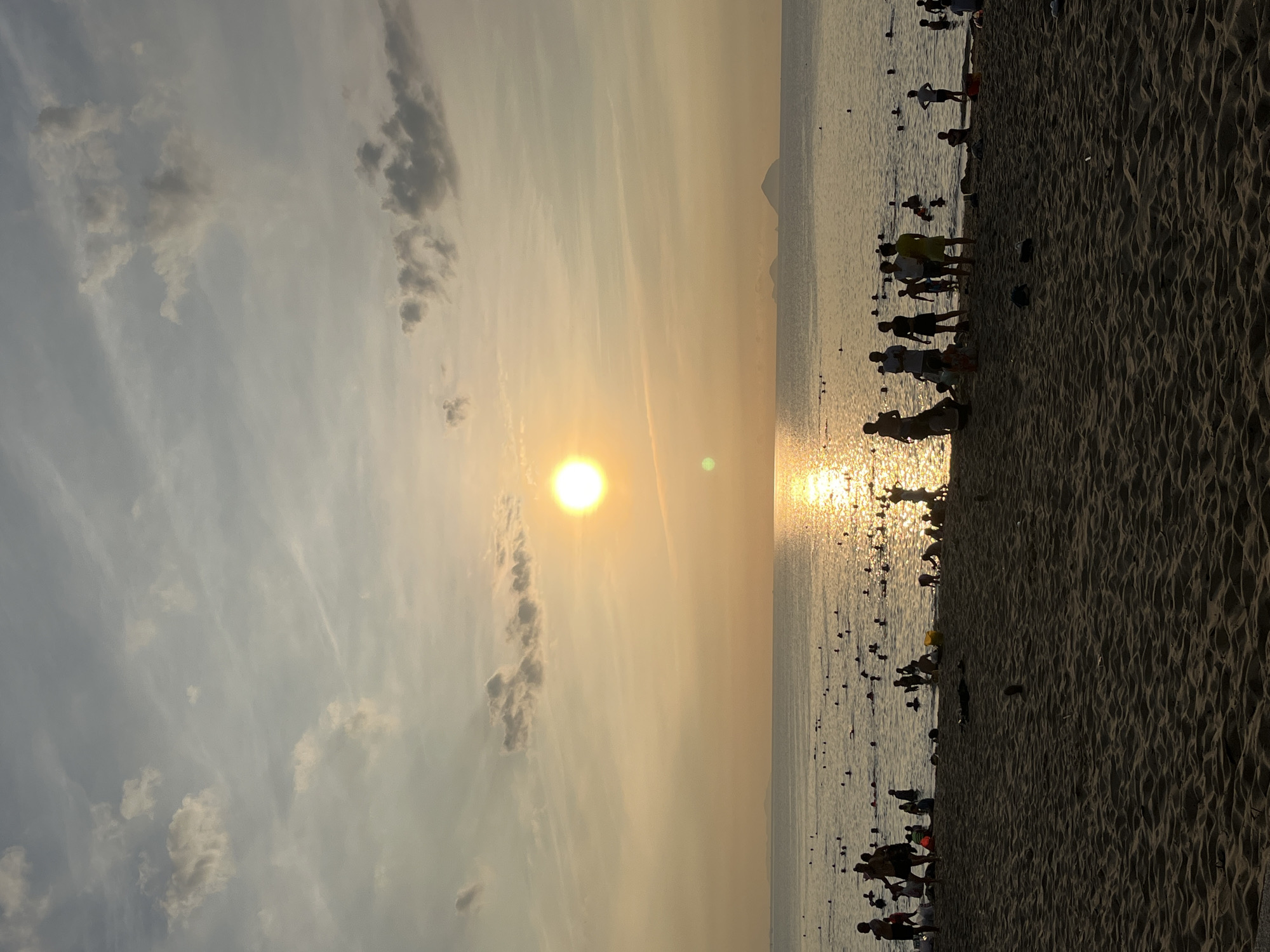 The image shows a beach scene at sunset in Nha Trang Ward, Vietnam. The sun is positioned in the sky, casting a golden glow over the water and sand. Silhouettes of people are visible along the shoreline, some walking and some standing near the water's edge. The beach is sandy with ripples. The sky is cloudy. In the distance, there are hints of a mountain range. The water has buoys.