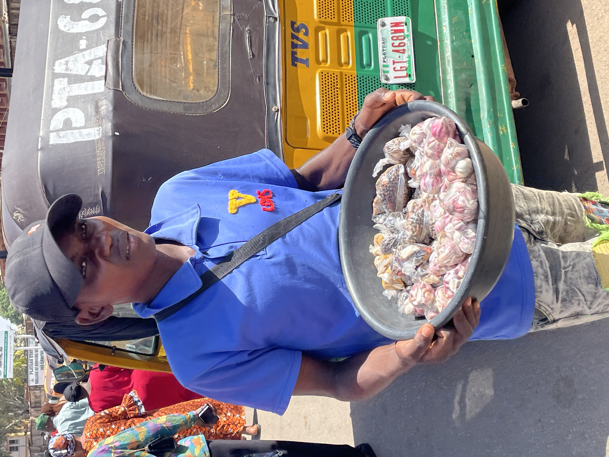 The image captures a bustling street scene in Jos, Plateau State, Nigeria, under bright daylight. A young man, likely a street vendor, is the central subject, looking towards the viewer with a neutral expression. He wears a blue short-sleeved shirt with "Y&S" embroidered in yellow and red, a black cap, and a black shoulder strap. He holds a large, dark basin filled with various items for sale, predominantly individual cloves of pinkish-white garlic neatly packaged in small clear plastic bags, alongside other dried goods or spices in similar packaging.
In the background, a yellow and green Keke Napep (auto-rickshaw) is visible, bearing the "TVS" brand name and a Nigerian license plate displaying "PLATEAU LGT-468WN." The Keke Napep's black canopy also has partially visible white lettering that appears to read "PTA-6". On the far left, a street sign is partially visible, showing "PLATEAU STAT COLLEGE OF NURSING SE JOS CAMPUS," which further confirms the location as Jos. Other people, including one in a distinctive orange and floral patterned outfit, are seen in the background. The clear shadows indicate a sunny day, highlighting the outdoor setting of this commercial activity.