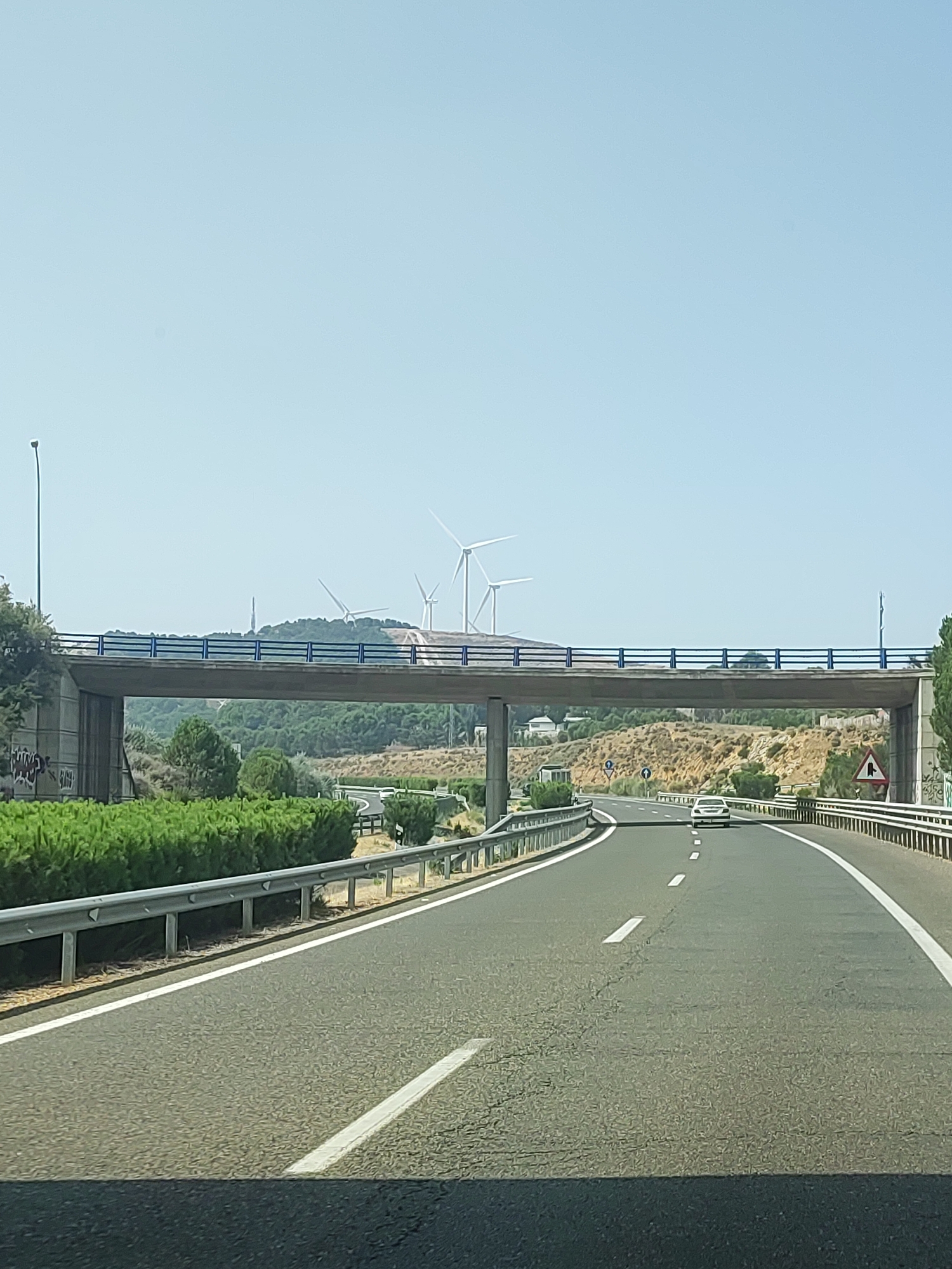 A bright, clear day with a pale blue sky. A highway stretches forward, curving slightly to the right. A white car is in the distance, traveling on the highway. A concrete overpass with blue railings crosses the highway. On the far side of the highway, there are wind turbines and a communications tower on a hillside. The area is landscaped with green bushes and dry, rocky terrain. The highway has white lane markings and metal guardrails on both sides. There is some graffiti on the left side of the overpass abutment. The image was taken from inside a vehicle, likely looking forward from the passenger seat.