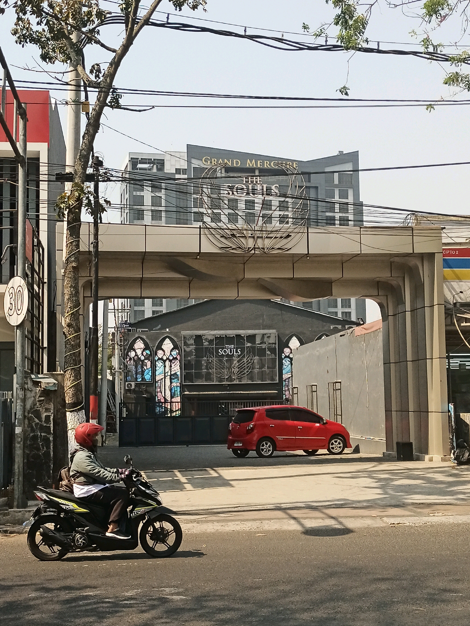 The image captures a street scene in Malang, Indonesia, specifically in front of the Grand Mercure hotel. A man wearing a red helmet is riding a black motorcycle on the road. He is wearing a gray jacket over a white shirt and dark pants, with a backpack on his back. A red car is parked on the side of the road near the hotel entrance. The hotel building is visible in the background, with its prominent "GRAND MERCURE THE SOULS" signage. The architecture features modern design elements, with large glass windows and clean lines. The foreground shows a tree with sparse leaves and utility poles with numerous electrical wires crisscrossing the sky. A speed limit sign indicating "30" is also visible on the left side of the image. The overall setting suggests a warm, sunny day in an urban environment.