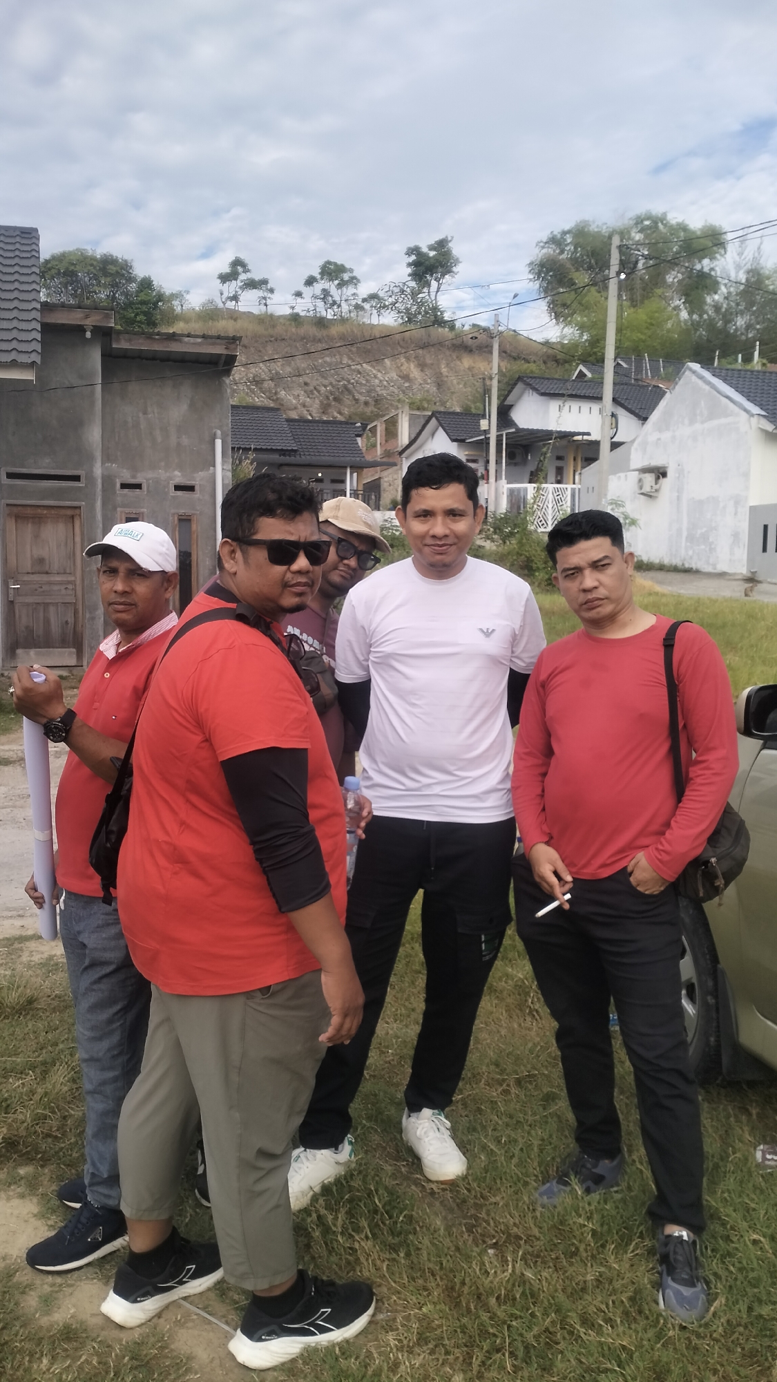 Five men stand in front of a cluster of houses under a cloudy sky, possibly in the city of Lhokseumawe, Indonesia. The man on the far left wears a red polo shirt and a white cap, holding a rolled-up document. Next to him, a man in a red t-shirt and sunglasses looks towards the camera. Behind him, a man in a maroon shirt and a tan cap is partially visible. The center of the group features a man in a white t-shirt and black pants, holding a bottle. To the right, another man in a red long-sleeved shirt and black pants holds a cigarette. The background shows houses with dark tiled roofs and some vegetation on a hillside. The ground is covered in grass.