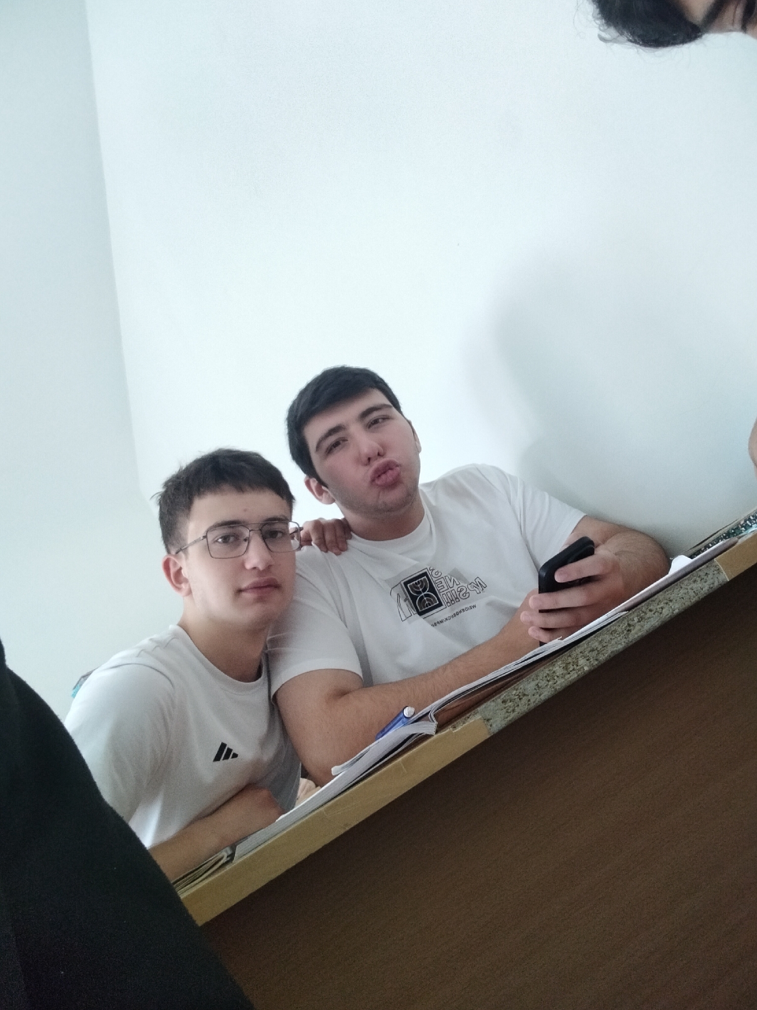 Two young men are seated at a desk with papers and a pen. The man on the left wears glasses and an Adidas t-shirt. The man on the right has a smartphone in his hand and is making a kissing face. They are in Yerevan, Armenia.