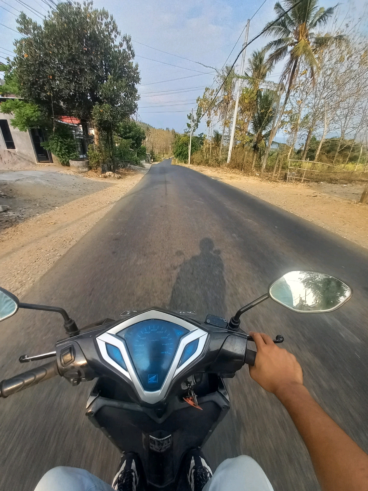 This is a first-person POV image of a person riding a scooter on a paved road in the city of Kulurejo, Indonesia. The road is flanked by a dirt path on the left and vegetation and trees on the right. In the distance, the road curves and appears to lead into a rural or semi-rural area with more trees and structures visible. A shadow of the rider and scooter is cast on the road. The scooter's dashboard with a speedometer is visible in the foreground. The sky is clear with a few clouds. The scene depicts a typical daytime commute or travel in the Indonesian countryside.