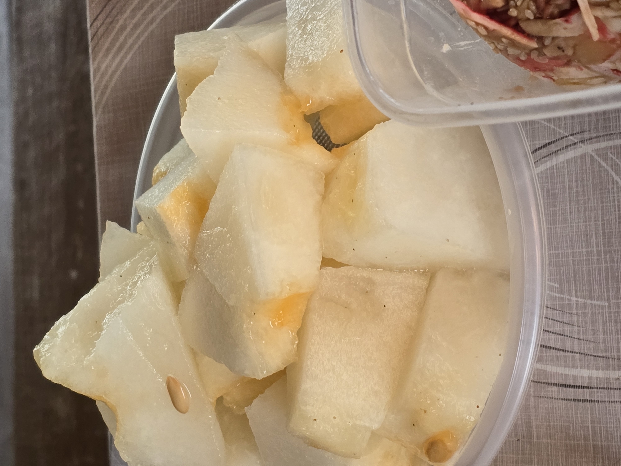 A close-up, overhead shot shows cubed melon in a clear plastic container, some pieces are triangular and some are cuboid. The melon is a pale yellow with some orange tinges visible. There are several seeds visible, one prominent oval-shaped seed is in the upper left quadrant of the image. The container is placed on a surface with a light-colored, woven pattern. To the right of the melon container, another clear plastic container is visible, containing a portion of a food item with red and white stripes and sprinkled with sesame seeds. The background consists of a textured, dark brown surface. The scene is indoors, with no discernable location within the city of Maksimiha, Russia.