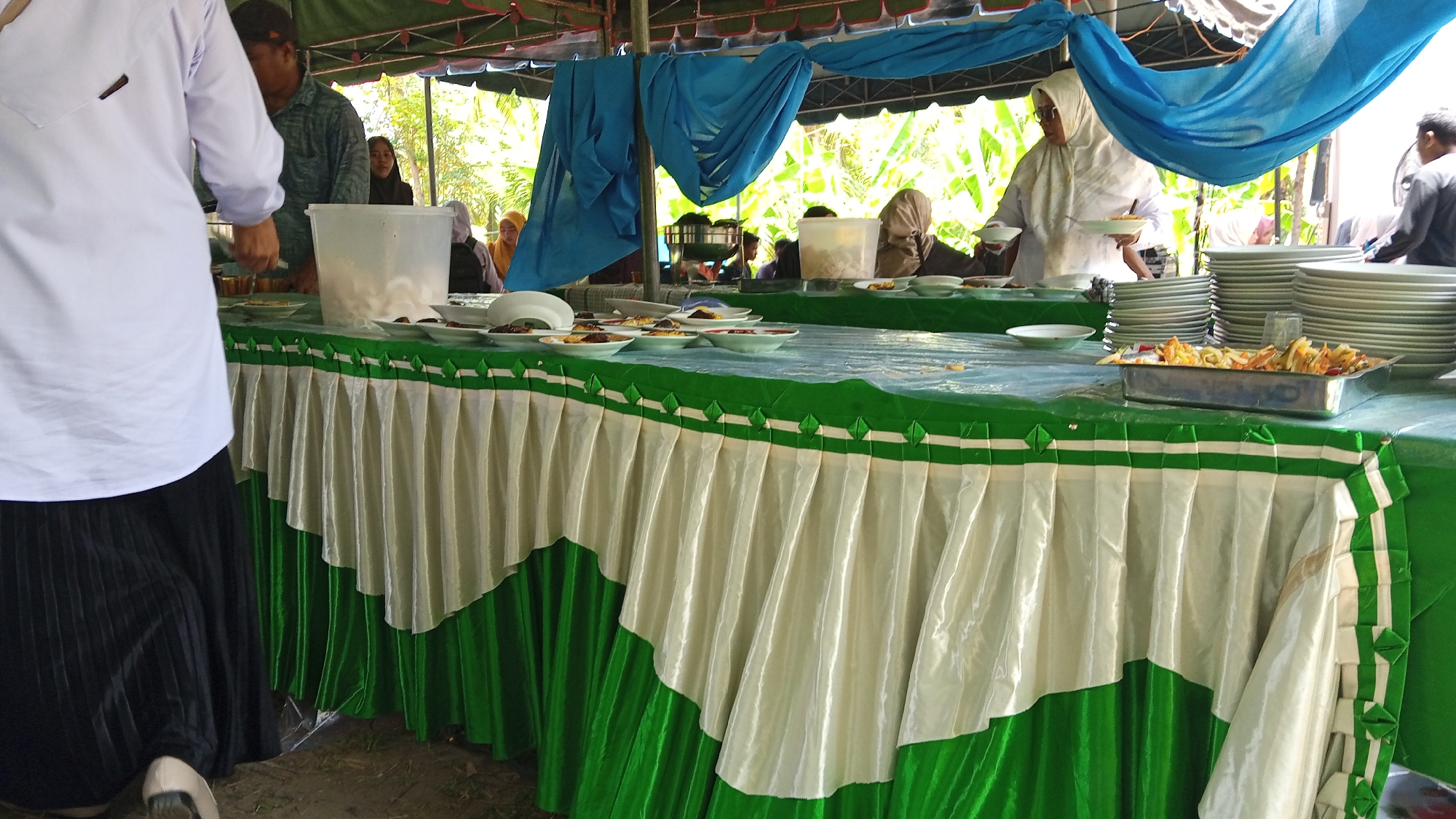 The image depicts an outdoor event, likely a catering setup, under a tent with blue and green fabric draping. A long table is covered with a green tablecloth and adorned with a draped skirting of cream and green fabric. Several stacks of white plates are visible on the table, along with serving dishes containing food. Some individuals, partially visible, are either attending to the food or are guests. One person in the foreground is wearing a white long-sleeved shirt and a black pleated skirt. The background shows foliage, suggesting a location in Blangrheuem, Indonesia, possibly at a community gathering or a celebration.
