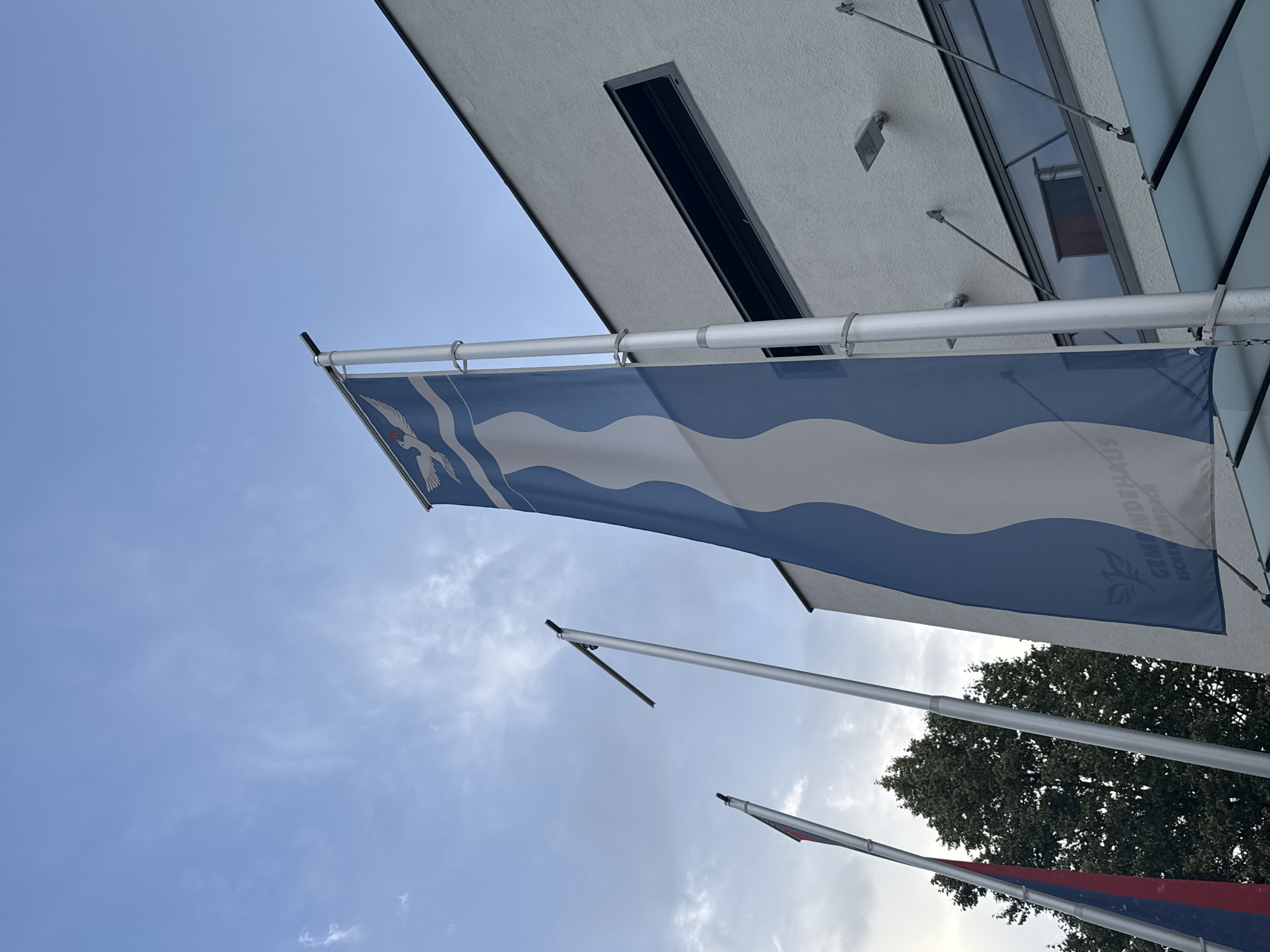 The image shows a low-angle view of a white building with a dark rectangular window, likely in Nendeln, Liechtenstein, as indicated by the text on the blue and white flag. Three flagpoles are visible. The largest flag, which is blue and white with a wavy pattern and a bird emblem at the top, hangs prominently in the foreground. Text on the flag reads "GEMEINDEHAUS" and "NESCHEN NENDELN". To the left of this flag, two other flags are partially visible. One is a tricolor flag with red and blue stripes and a white section. Behind the flags, a large tree with dark green foliage is visible against a sky with scattered clouds. The building's facade is textured white, and a light fixture is mounted on the exterior wall near the window. A section of a glass-covered awning extends from the building's entrance area.
