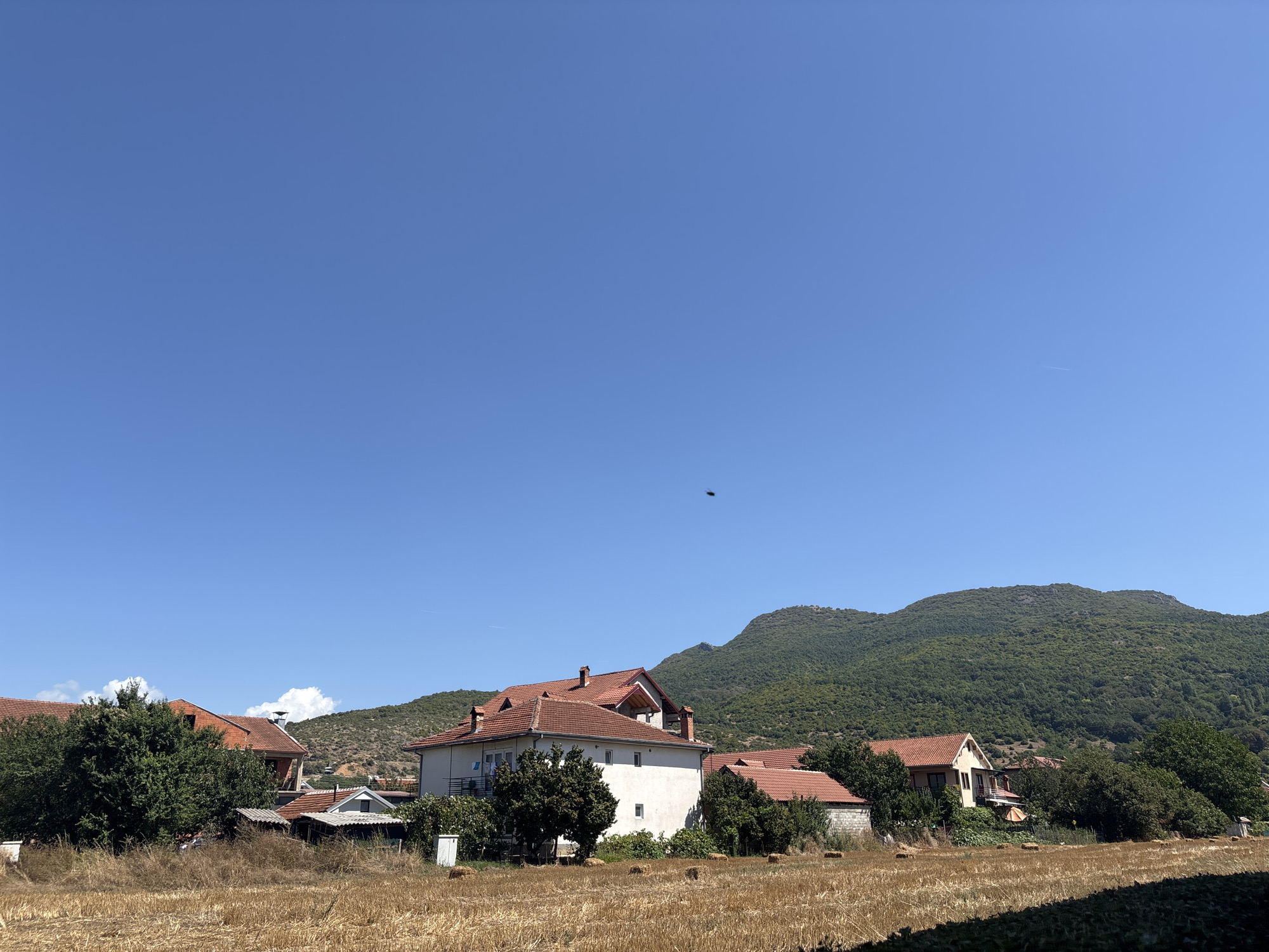 The image depicts a rural scene in the vicinity of Leskoec, North Macedonia, on a clear, sunny day. In the foreground, a field of dry, harvested hay is visible, with several rectangular hay bales scattered across it. Behind the field, a cluster of houses with red-tiled roofs are nestled amongst lush green trees. The architecture of the houses appears to be traditional, with some featuring balconies and chimneys. In the background, rolling green hills covered in dense vegetation form a natural backdrop. The sky is a vast expanse of clear blue, with a single small dark object, possibly a bird or insect, visible in the upper portion of the frame. The overall impression is one of a peaceful, rural landscape under a bright sky.