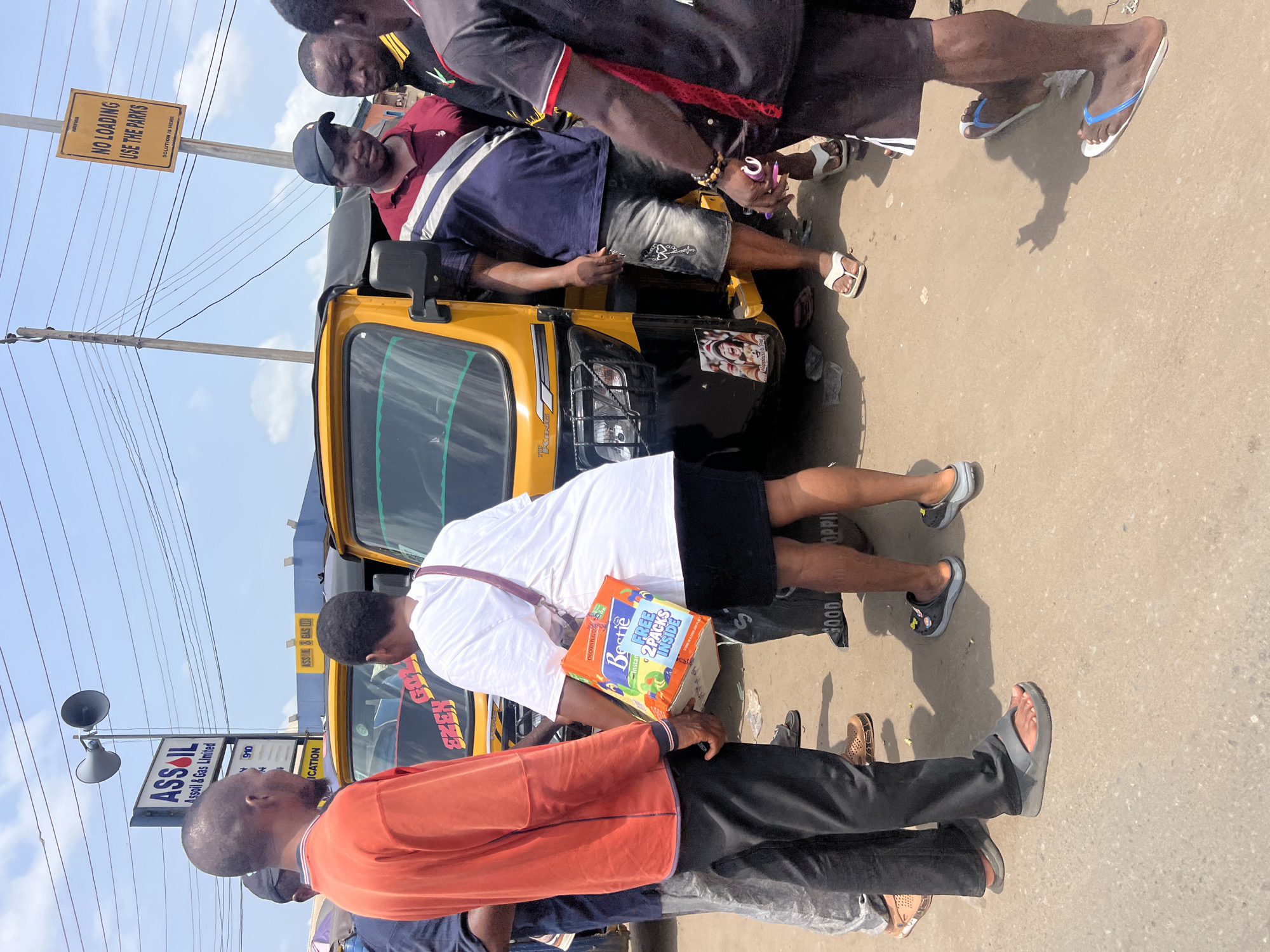 The image shows a street scene in Onitsha, Nigeria. A yellow three-wheeled vehicle, possibly a tuk-tuk, is visible with several people standing nearby. An ASSOIL gas station sign is in the background, as well as a sign that says, "NO LOADING USE THE PARKS, SOLUTION IS HERE." A person in a white shirt is carrying a box labeled "Bestie Instant Noodles FREE 2 PACKS INSIDE." The image captures a typical urban environment with everyday activities taking place.