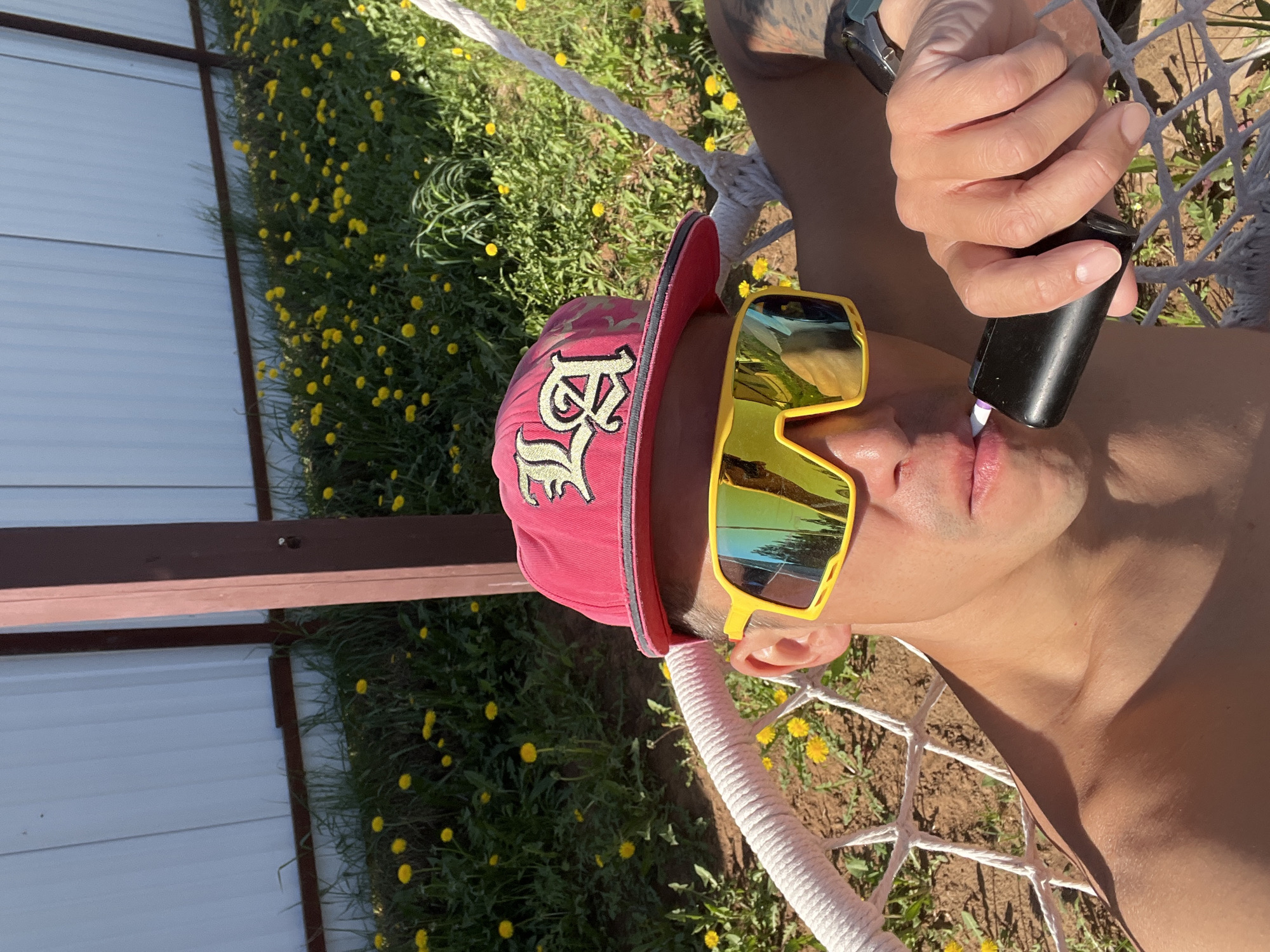 A man is reclining in a hammock, facing towards the camera. He is wearing a red baseball cap with a light-colored embroidered design, and yellow-framed sunglasses with mirrored lenses. He is holding a black vape pen to his lips. The surrounding area features a white building with a dark brown beam, along with green vegetation and yellow flowers. The location could possibly be Chaikovsky, Russia.