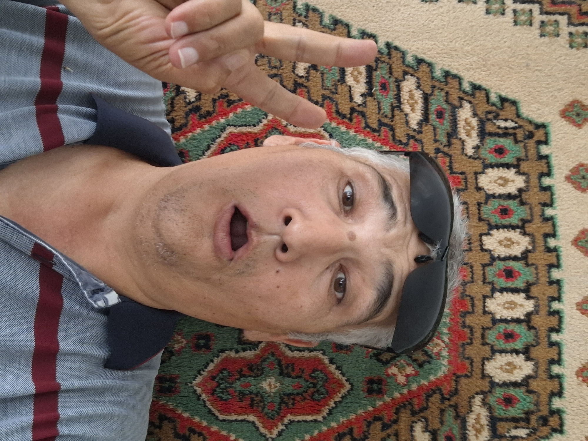 A man is lying down on a patterned carpet, looking up at the camera. He is wearing sunglasses on his head and a striped polo shirt. His mouth is open, and he is holding up two fingers in a peace sign with his left hand. The carpet has a repeating geometric pattern in shades of beige, green, red, and black. The man's skin tone is fair, and he has graying hair. The lighting is bright, suggesting it is daytime. No specific location in Almaty, Kazakhstan can be determined from the image.