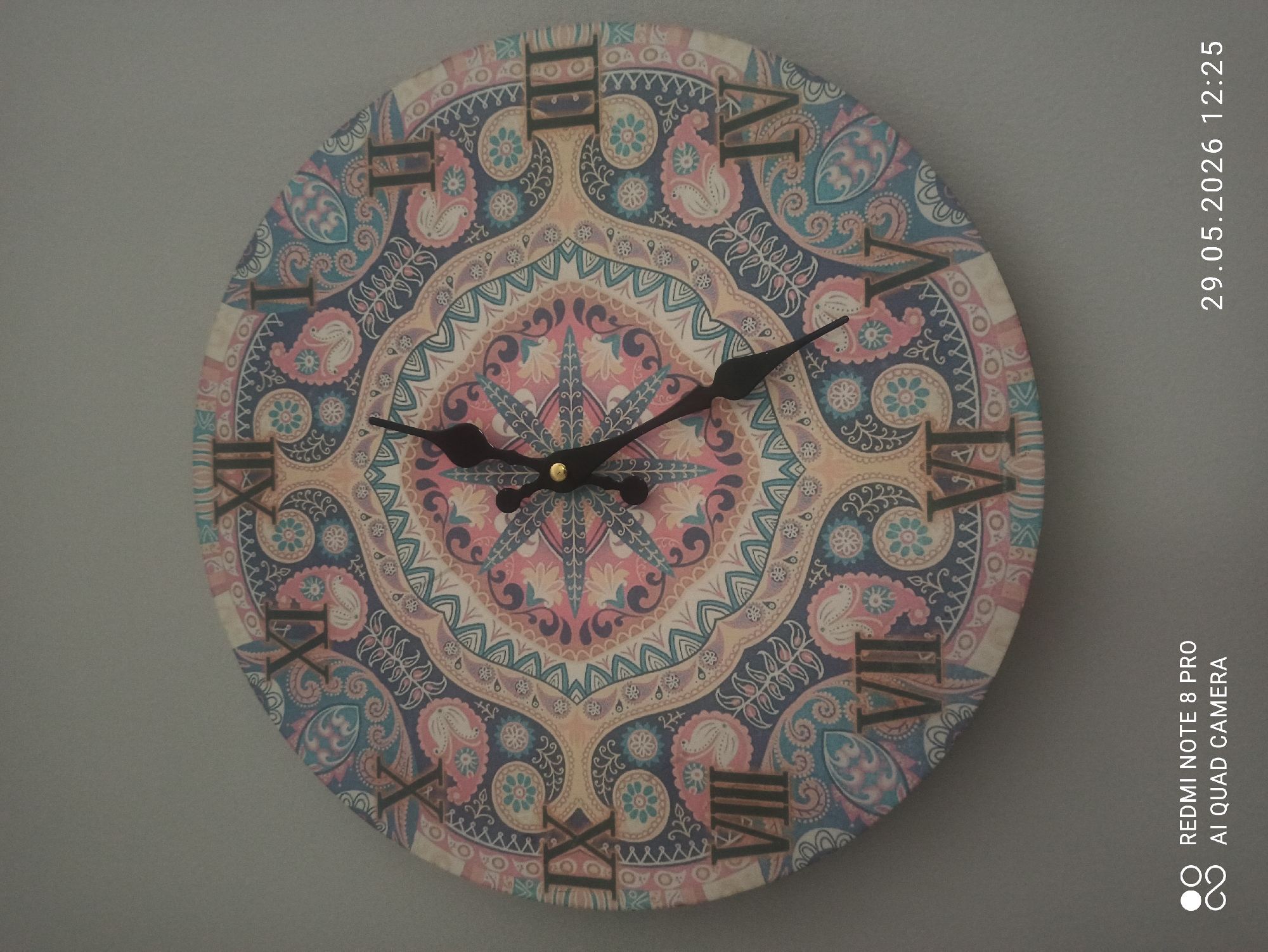 Here is a forensic description of the image:
The image shows a close-up of a round, decorative wall clock hanging on a light gray wall. The clock face is adorned with a complex, colorful paisley-like pattern in shades of blue, pink, peach, and navy. Black Roman numerals indicate the hours. The clock hands are black, with the minute hand pointing towards the 2 and the hour hand around the 9.
The location is Unknown, Unknown. The presence of the timestamp "29.05.2026 12:25" and "REDMI NOTE 8 PRO AI QUAD CAMERA" suggests a digital image captured by a mobile device camera, possibly in an indoor environment.