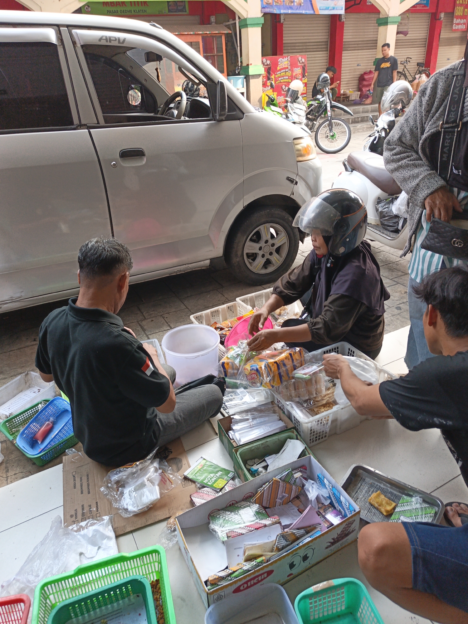 The image depicts a street scene in Klaten, Indonesia, likely a market or roadside stall. In the foreground, three individuals are engaged in what appears to be a transaction or organization of goods. A man in a black polo shirt, with a small Indonesian flag patch on his left sleeve, sits on the ground facing away from the camera, interacting with a white bucket and plastic containers. To his right, a woman wearing a black hijab and a helmet is seated, actively sorting through various packaged food items and small goods displayed in white plastic baskets. Her hands are engaged with the items. Another person, partially visible from the back and wearing a black t-shirt and blue shorts, is seated to the right, also seemingly involved with the items.
Surrounding the individuals are numerous baskets and boxes filled with a variety of products, including packaged snacks, small items in clear plastic, and what look like small booklets or cards. The ground is paved with square tiles. To the left of the scene, a silver car is parked, partially obscuring the background. Behind the individuals, a market stall with red pillars and a closed shutter can be seen. Further in the background, several motorcycles are parked, and other individuals can be observed, one standing and another on a bicycle. The overall impression is one of a busy commercial area where goods are being sold or managed outdoors.