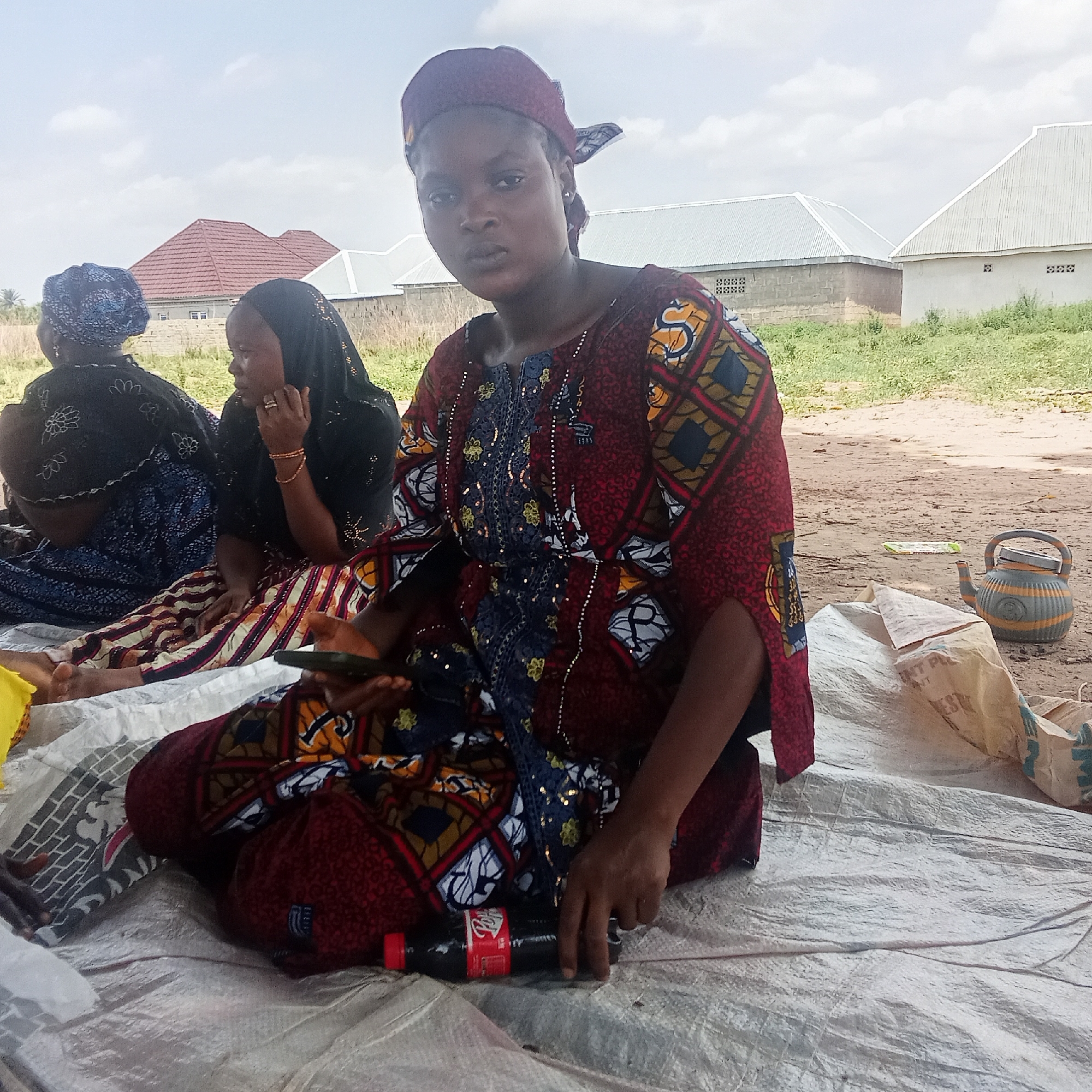The image depicts a group of three women seated on a white tarp, likely in an outdoor setting in Doma, Nigeria.
The woman in the foreground wears a patterned, maroon-colored dress with a matching head covering. She is holding a black smartphone in her left hand and a Coca-Cola bottle is on the tarp near her right hand.
Two other women are seated behind her to the left. One is wearing a patterned blue headscarf, and the other wears a black headscarf.
In the background, buildings are visible, including houses with roofs of different colors. A teapot is visible on the ground near the edge of the tarp. The ground appears to be sandy. The sky is overcast.