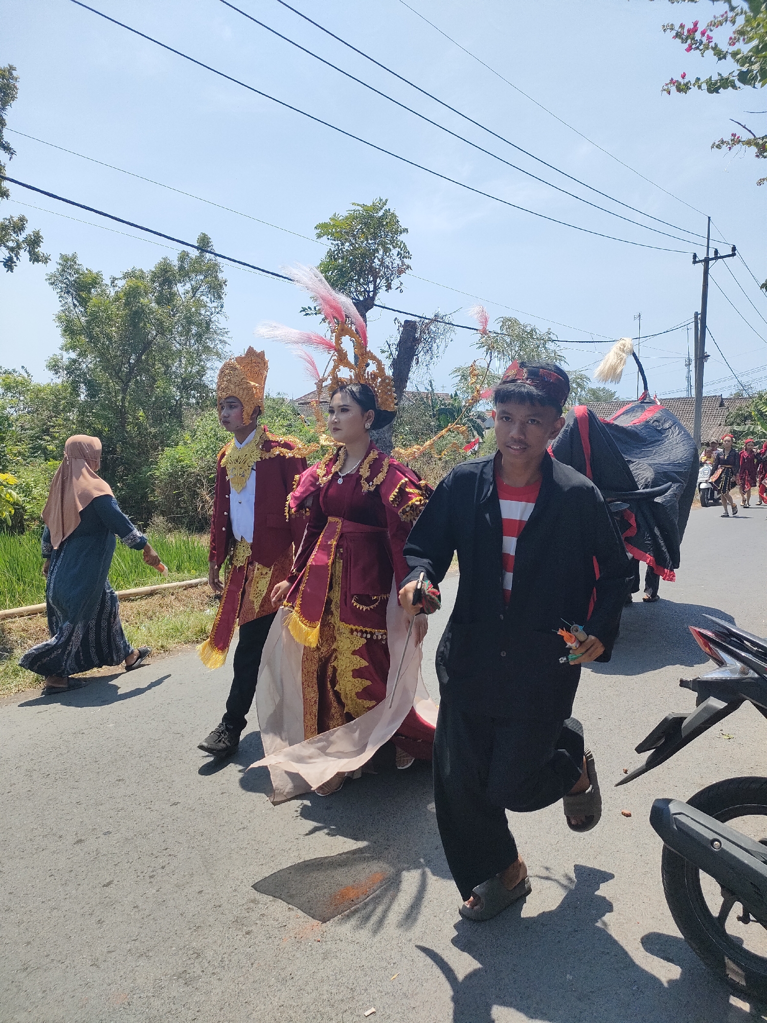 This image captures a street scene in Tongaswetan, Indonesia, under a clear, bright sky. A procession is underway, with several individuals in traditional attire. In the foreground, a young man in a black jacket and a striped red and white shirt runs forward, holding what appear to be tools or props. Behind him, a woman and a man are dressed in ornate, red and gold traditional garments, with elaborate headdresses. The woman wears a red velvet dress with intricate gold embroidery and a sheer veil. The man is adorned in a red jacket with gold trim and a golden headdress. To the left, a woman in a dark blue hijab and dress walks away from the camera. Further in the background, a large, dark fabric-covered structure, possibly a costume or float, is visible, with white tufts resembling a tail. Other people and a motorcycle are also present, suggesting a festive or ceremonial event is taking place. The environment features a paved road, green foliage, and trees lining the sides, with houses visible in the distance. Power lines are strung across the sky.