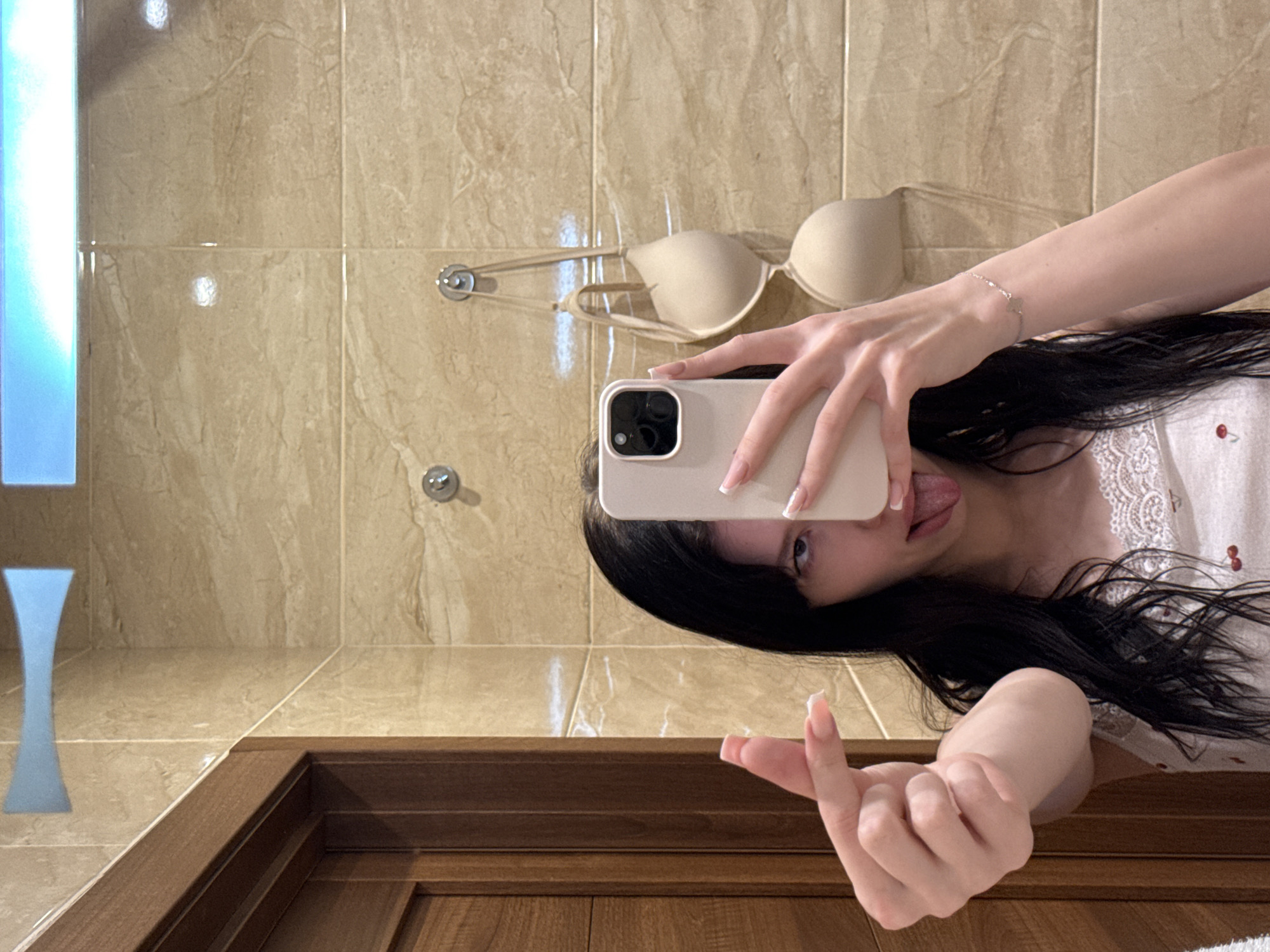 A young woman with long, dark hair is taking a selfie in a bathroom. She is wearing a white top with red cherry embroidery and white lace trim. She is sticking her tongue out and making a heart shape with her fingers. Her phone is held up to take the photo. In the background, a beige bra is hanging on a hook on the tiled wall. A wooden door is visible on the left side of the image. The scene is in a bathroom in Khimki, Russia.