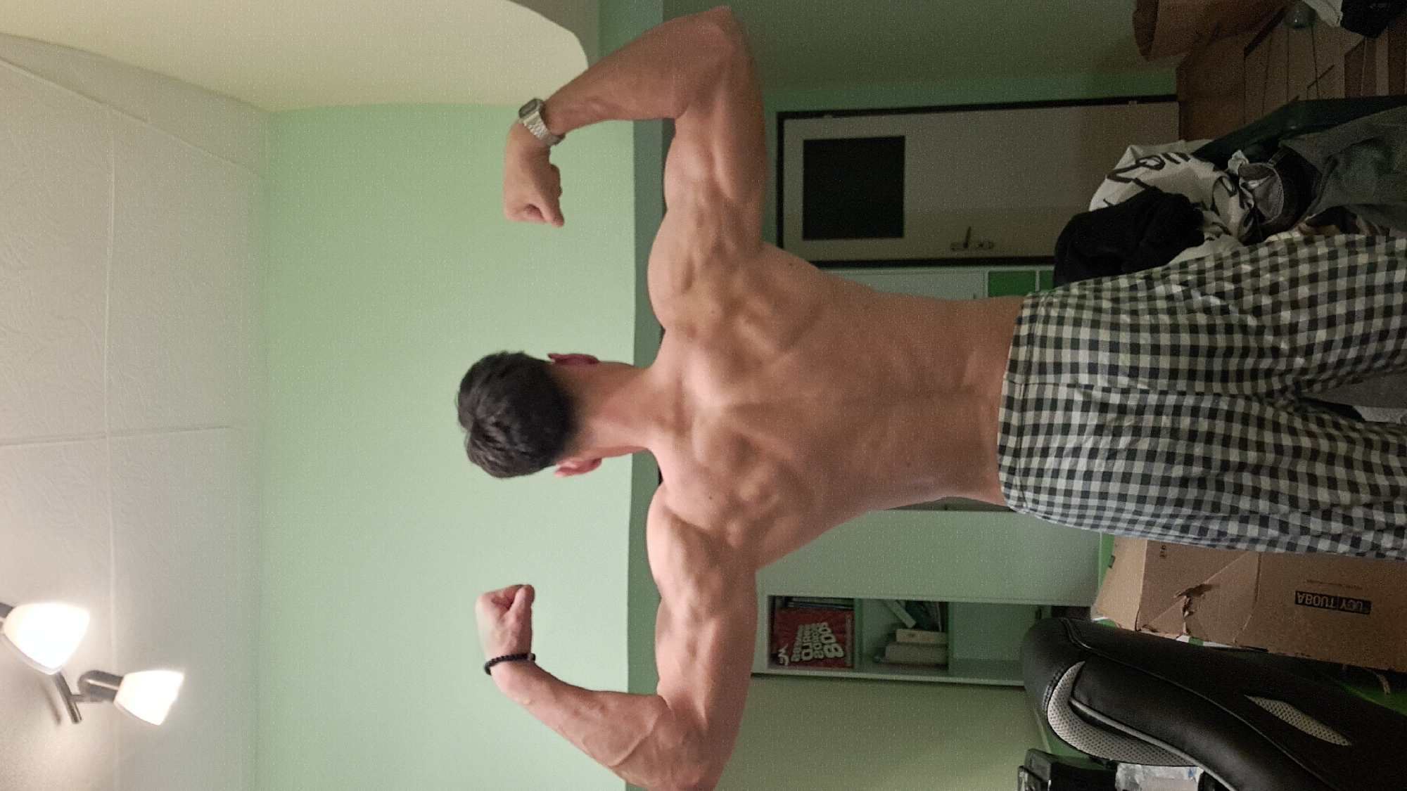 A muscular man with dark hair is posing shirtless, displaying his back and biceps. He is wearing black and white checkered pants. He has a watch on his left wrist and a bracelet on his right wrist. The background includes a light green wall, a white door with a dark square inset, a shelf with books and a box labeled "UCYT TUOBA", a black chair, and a ceiling with a light fixture. There is no discernible information about the location being in the city of Burgas, Bulgaria.