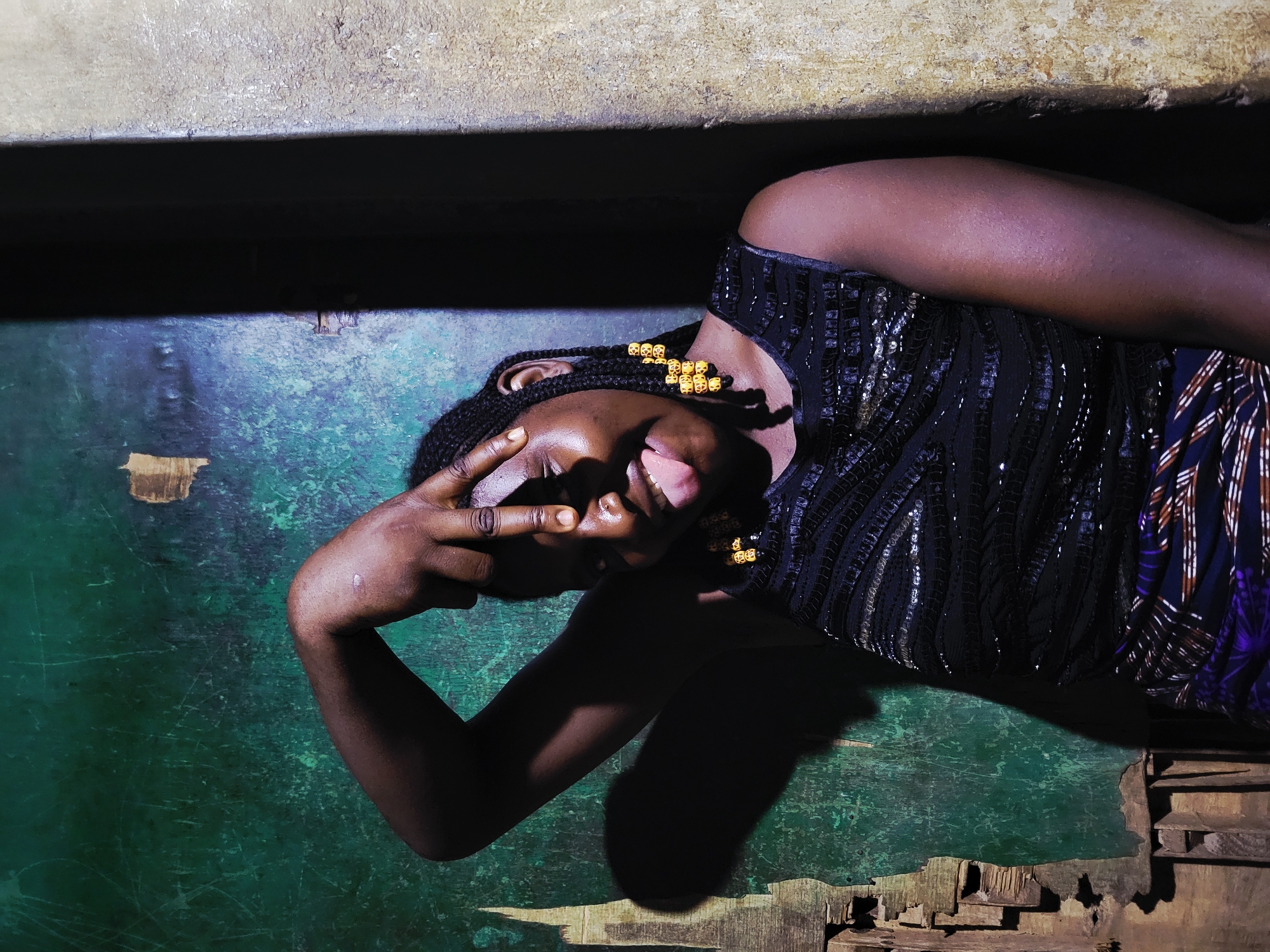 A horizontal eye-level close-up shot shows a person with dark skin, possibly female, lying on a blue-green cracked and scratched surface under a horizontal grey concrete ledge. The person's head is tilted, facing the camera, with their hand covering their face, and a portion of their tongue visible. The person is wearing a dark top with vertical sequin-like embellishments. They also have braided hair with gold-colored beads. Behind the person, partially obscured, are stacked, weathered wooden boards. The location, Jos, Nigeria, is not readily evident from the image alone.