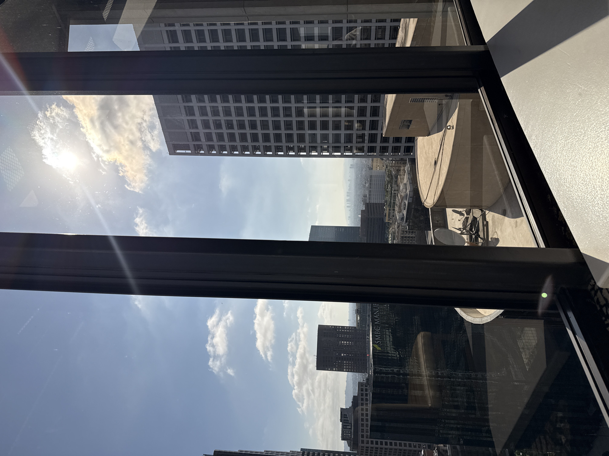 The image shows a view of the Los Angeles skyline through a window. The sun is shining brightly, casting rays of light and creating lens flare. Several tall buildings are visible, including a grid-patterned skyscraper on the right and a darker, square-shaped building with many windows in the center. In the foreground, there is a curved structure and what appears to be a rooftop with some equipment. Reflections of the interior of the room are visible on the glass. The sky is blue with scattered clouds. The overall impression is of a sunny day in a densely populated urban environment. The specific location is likely a high-rise office building in downtown Los Angeles, given the density and style of the architecture.