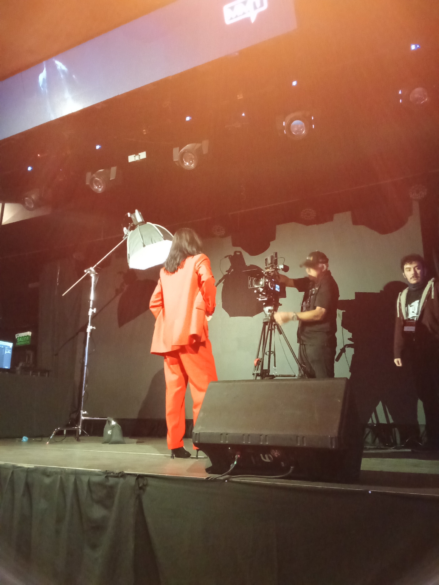 In this medium shot, three individuals are on a stage, presumably during an event in Santiago, Chile. The person in the foreground, viewed from behind, is dressed in a vibrant, formal red suit and black heels, with long dark hair. To the left of this individual, a professional lighting setup with a softbox is visible.
Center stage, a person wearing a black t-shirt, black pants, and a baseball cap operates a professional video camera mounted on a tripod. Their focus is on the person in the red suit. To the right, another person, dressed in a black hoodie and a dark shirt with a white graphic, stands facing towards the camera operator and the main subject.
The background features a large screen displaying abstract imagery and a logo that appears to read "MAXI". Above the screen, stage lighting fixtures are visible. The stage itself is covered with a black curtain. On the left side of the stage, a monitor with the word "SALIDA" (Spanish for "Exit") is mounted on a stand, suggesting this may be a backstage area or a backstage interview setup. The lighting is dramatic, with some lens flare present, indicating artificial stage lighting. The overall mood appears to be professional and focused on production.