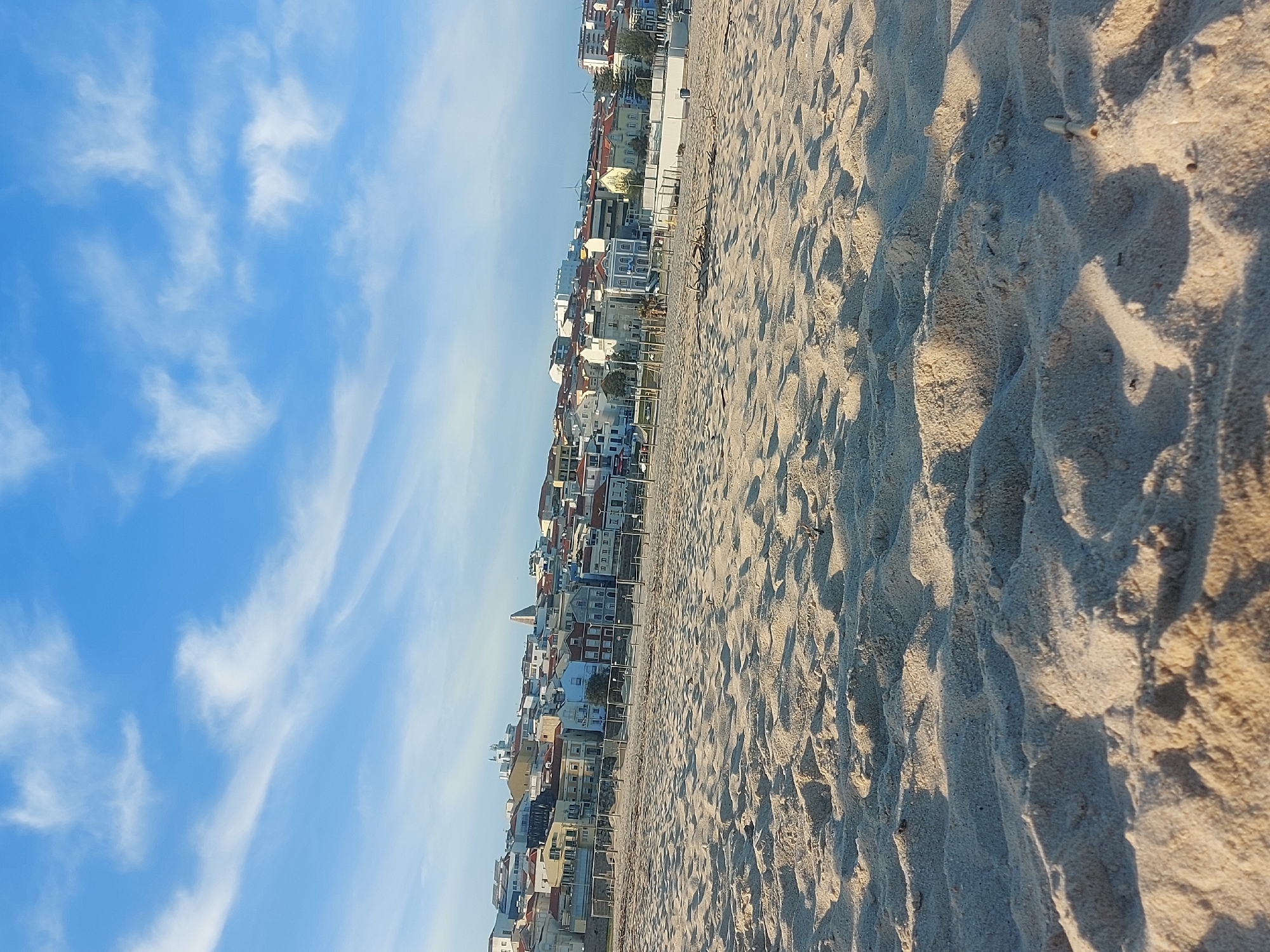 The image captures a view of Figueira da Foz, Portugal, specifically the beach area. The scene is divided between a clear blue sky with elongated white clouds and a sandy beach. The sand is textured with shadows, suggesting the presence of undulations or footprints. Along the horizon line, a series of buildings form the urban edge, transitioning from the beach to the city. The perspective is somewhat tilted. There are no people visible in the image. The overall environment suggests a sunny day at the beach with the city as a backdrop.