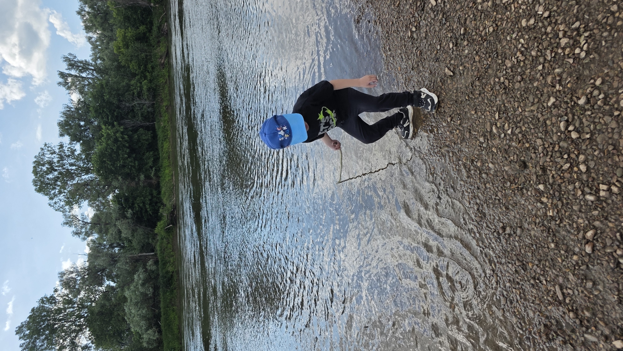 A young boy wearing a blue baseball cap and a black t-shirt with a graphic print is standing on a pebble-strewn riverbank. He is holding a stick and appears to be poking it into the shallow water. The river is calm with ripples on its surface. The background features a dense line of green trees and a cloudy sky. The scene is set in an outdoor, natural environment. The location is identified as the city of Дарьино, Russia.
