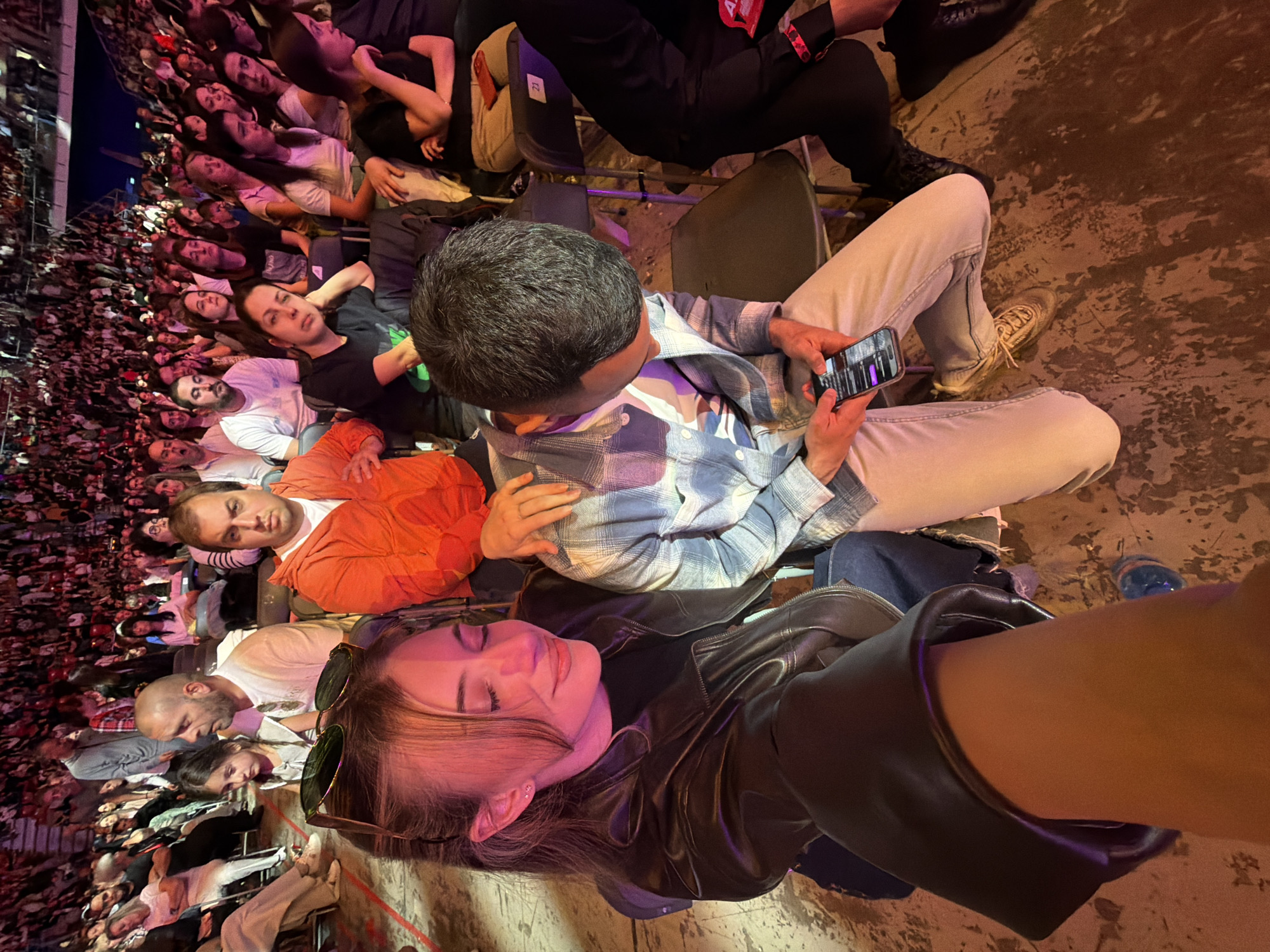 The image shows a crowded concert venue, possibly in Sofia, Bulgaria, although no specific landmarks are visible. A dense crowd of people fills the background. In the foreground, a woman with long brown hair and sunglasses resting on her forehead reclines, seemingly relaxed, wearing a black leather jacket. Adjacent to her, a man wearing a blue and white plaid shirt is looking at his mobile phone. The man is wearing light colored jeans and sneakers. The floor appears to be concrete with some staining. The lighting suggests an indoor venue with mixed artificial light sources. The perspective suggests the photo was taken from a high angle.