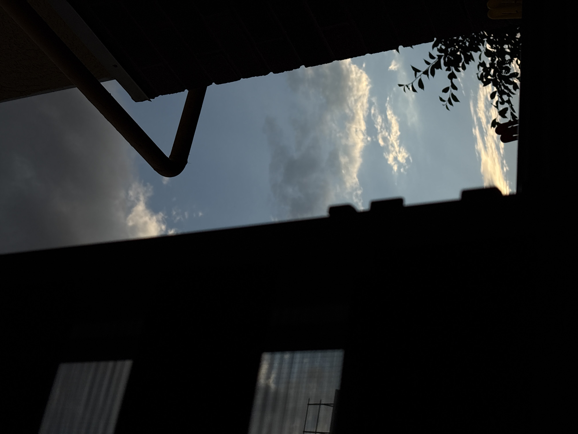 This media file depicts an outdoor scene viewed from a low angle, looking up towards the sky. The foreground is dominated by the dark silhouettes of structures, possibly a roofline and a window frame. One silhouetted structure features what appears to be a drainpipe extending diagonally.
The sky, visible through an opening in these structures, is a light blue with scattered clouds. Some clouds are white and fluffy, while others appear darker and more overcast, suggesting a varied weather pattern. The lighting indicates it is likely late afternoon or early evening, as the clouds on the right side of the sky are illuminated with a warm, golden hue. The presence of leaves and branches, also silhouetted against the sky in the upper right quadrant, suggests the scene is near trees or vegetation.
Below the sky and within the dark foreground, there are two rectangular openings. These openings show a blurry, ribbed texture, possibly indicating glass or plastic material with vertical lines. A faint, thin rectangular structure is visible within the right-hand opening, resembling a metal support or frame.
The overall mood is somewhat mysterious and introspective, with the contrast between the dark foreground and the bright sky creating a dramatic effect. There are no people or visible text in the image. The location context of Mikhaylovsk, Russia, does not provide specific visual cues within the image itself.