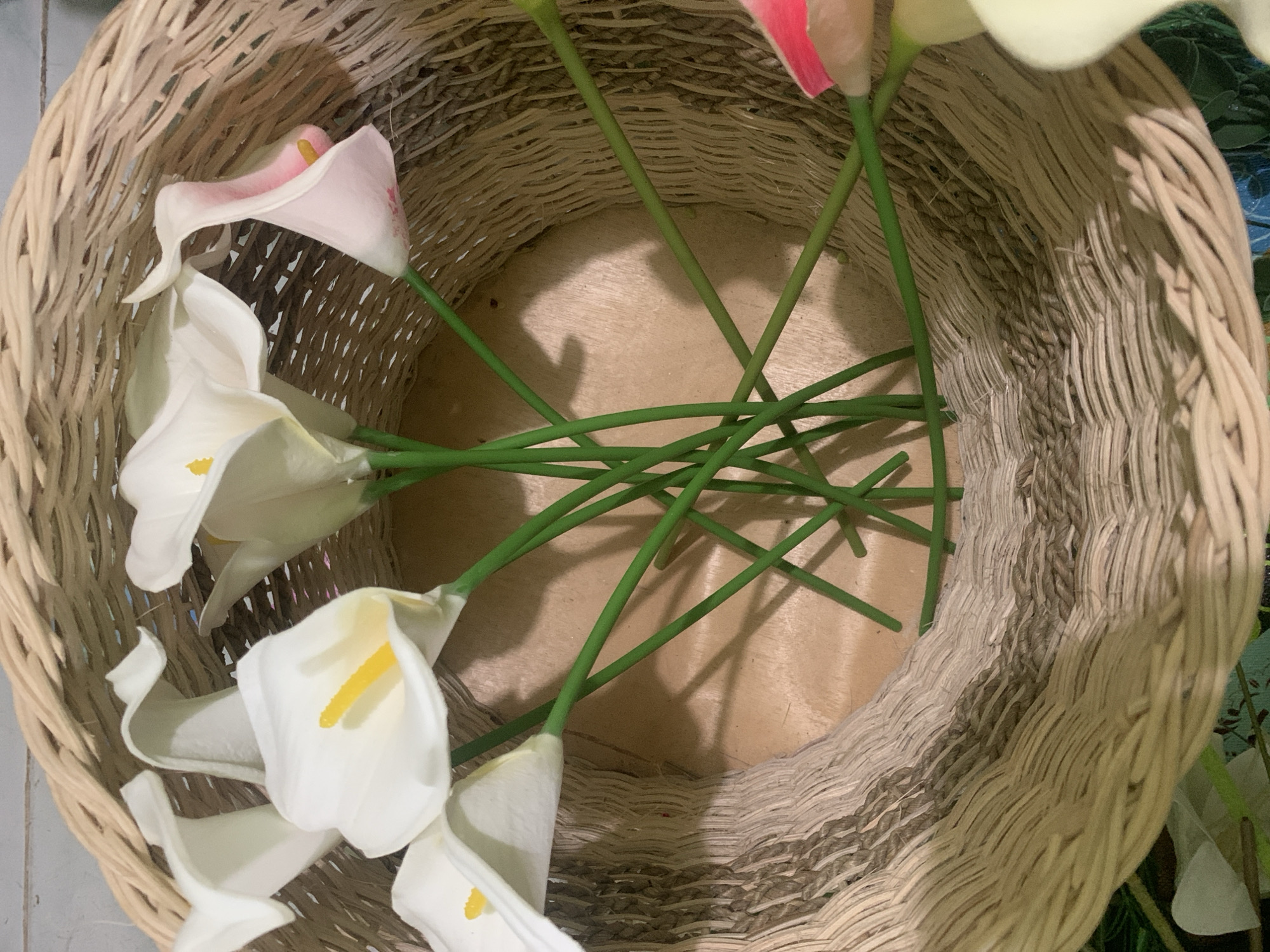 The image shows a close-up view of a woven basket containing several artificial calla lilies. The basket is made of light-colored wicker, with a textured pattern. Inside the basket, several white calla lilies with yellow centers are visible, along with one pink and white calla lily. The stems of the lilies are green and are arranged in a fanned-out manner. The interior of the basket appears to be made of a light-colored wood. The lighting suggests an indoor setting, possibly with natural light casting shadows. There are no people or text visible in the image. The overall impression is that of a decorative arrangement.