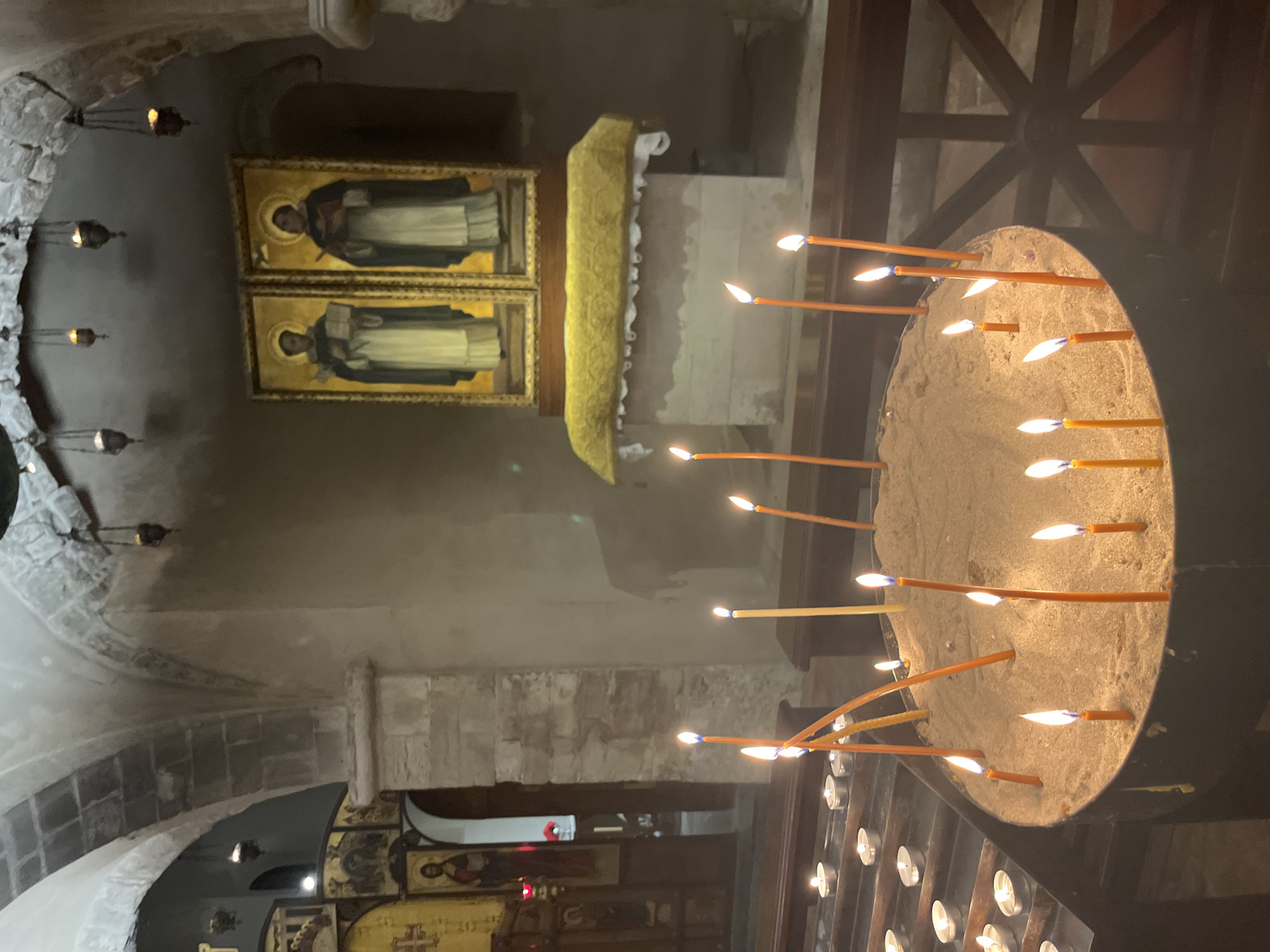 An indoor image captures a dimly lit interior of a church, likely in Bari, Italy, characterized by stone architecture and religious iconography. In the foreground, a large cylindrical metal container filled with sand holds numerous lit, thin orange candles. To the left, a tiered metal rack displays a row of smaller, lit white votive candles. In the background, two framed religious icons depicting robed figures with halos are displayed on a raised platform covered with a yellow cloth. The ceiling features stone arches with suspended, dimly lit lanterns. The overall atmosphere is one of solemnity and devotion.