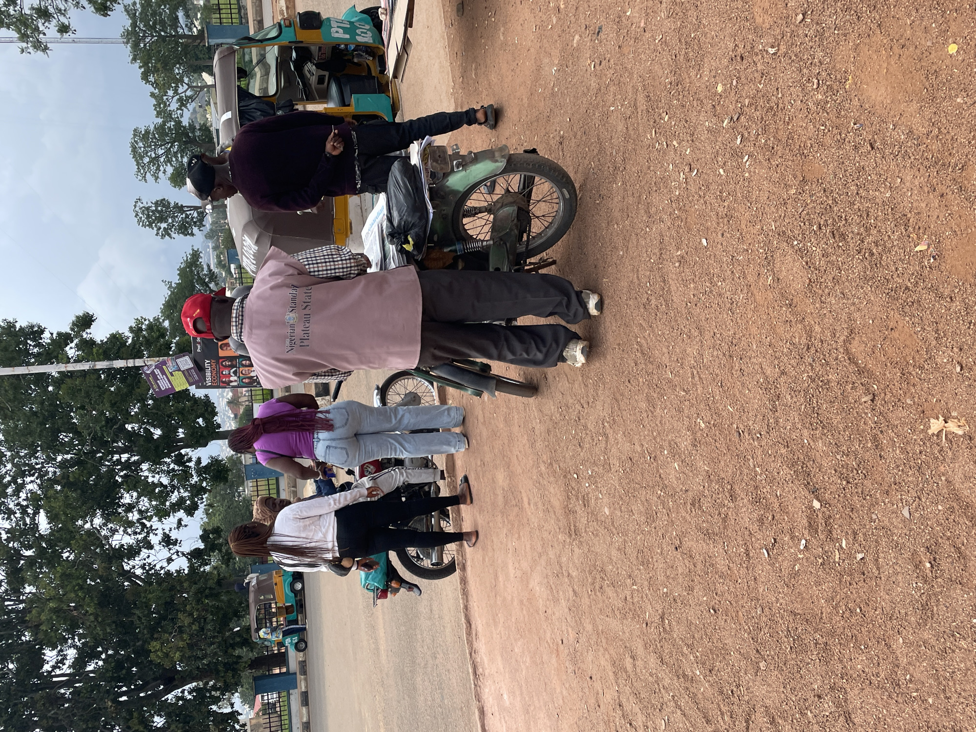 A group of people are standing around a motorcycle and a small yellow motorized tricycle on a dirt road. Two men, one wearing a dark purple sweater and a black cap, and another wearing a red cap and a pink shirt with text, are near the front of the scene. Behind them, two women are visible, one in a purple top and light blue jeans, and another in a white shirt and black pants. The motorcycle appears to be older and green, with some visible rust. The tricycle is brightly colored yellow with some blue accents and has "PT 100" written on it.
The overall scene suggests a casual street setting in a location with dusty, unpaved roads, consistent with Bar Jirgi Summa, Nigeria. The lighting suggests it is daytime, likely overcast or late afternoon given the soft shadows. The people appear to be casually interacting, with no obvious signs of distress or urgency.
Visible text includes "Nigerian Standard Plateau State" on the pink shirt and "PT 100" on the tricycle. There are also some less legible words on posters or signs in the background, including "VISIBILITY ECONOMY".