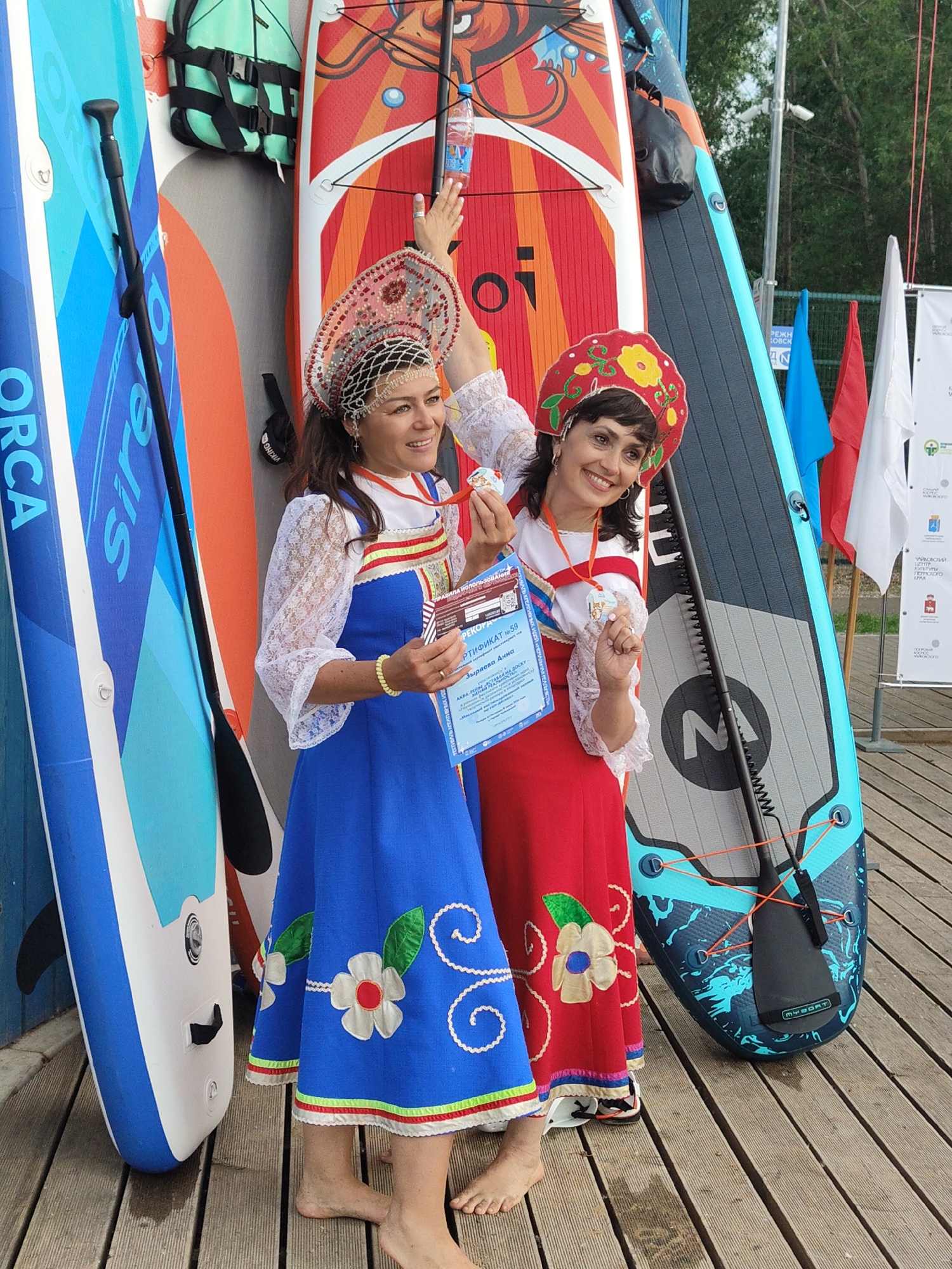 Two women, dressed in traditional Russian attire, are posing with paddleboards. The woman on the left wears a blue dress with white and red trim and floral appliques, and a jeweled headdress. She holds a certificate and a medal. The woman on the right wears a red dress with floral appliques and a red headscarf with floral embroidery. She also holds a medal. They are standing on a wooden deck next to several paddleboards, one of which has a "MYBOAT" logo. In the background, there are flags and trees, suggesting an outdoor event, possibly a competition or festival. The weather appears to be fair.