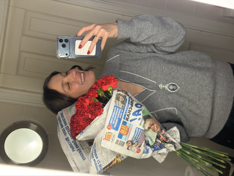 A woman smiles while holding a bouquet of red carnations wrapped in newspaper. She is taking a selfie with a phone that has a gray case and a pop-it accessory. She is wearing a gray sweater with white trim and a cameo brooch. The photo appears to be taken in a bathroom, as indicated by the wall-mounted light fixture and the corner of the countertop visible.