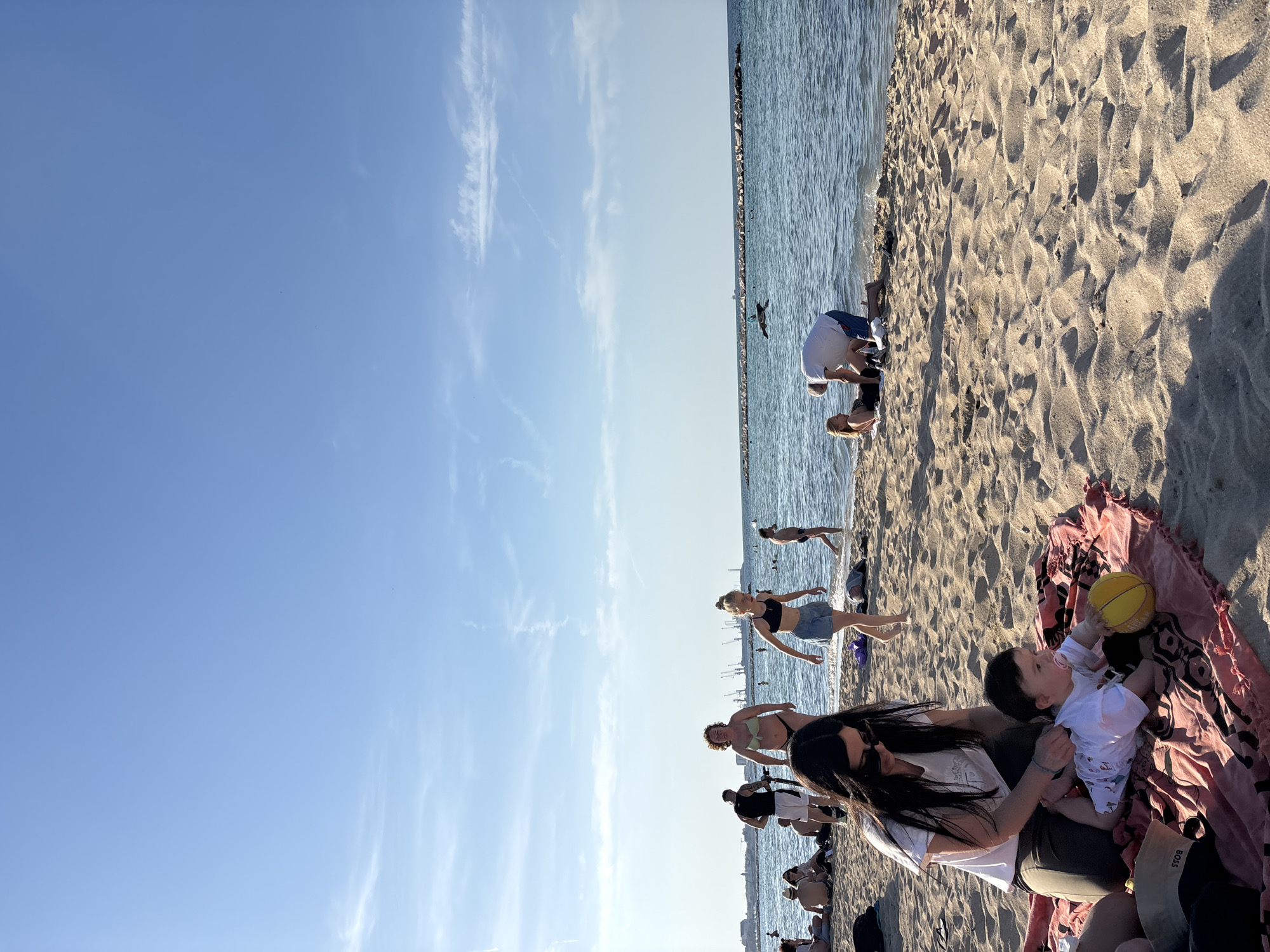 The image captures a lively beach scene under a clear blue sky with faint wispy clouds. In the foreground, on the sun-drenched sand, a woman with long dark hair, wearing sunglasses and a white t-shirt, looks down at a baby. The baby, dressed in a white patterned onesie, is seated on a pink and red patterned blanket and holding a yellow ball. A black bag with "BOSS" written on it is visible near the blanket.
Numerous other people are scattered along the beach and in the calm, blue water. Several individuals are seen wading or standing in the shallow parts of the water, including one person in a light-colored bikini top and another in a black sports bra and denim shorts. Further down the shore, a man in a white t-shirt is bent over, interacting with the sand, with another person close by. In the distance, a line of rocks, possibly a breakwater or jetty, extends into the water. The bright sunlight casts distinct shadows across the textured sand. The scene conveys a relaxed and enjoyable atmosphere, characteristic of a sunny day spent by the ocean.