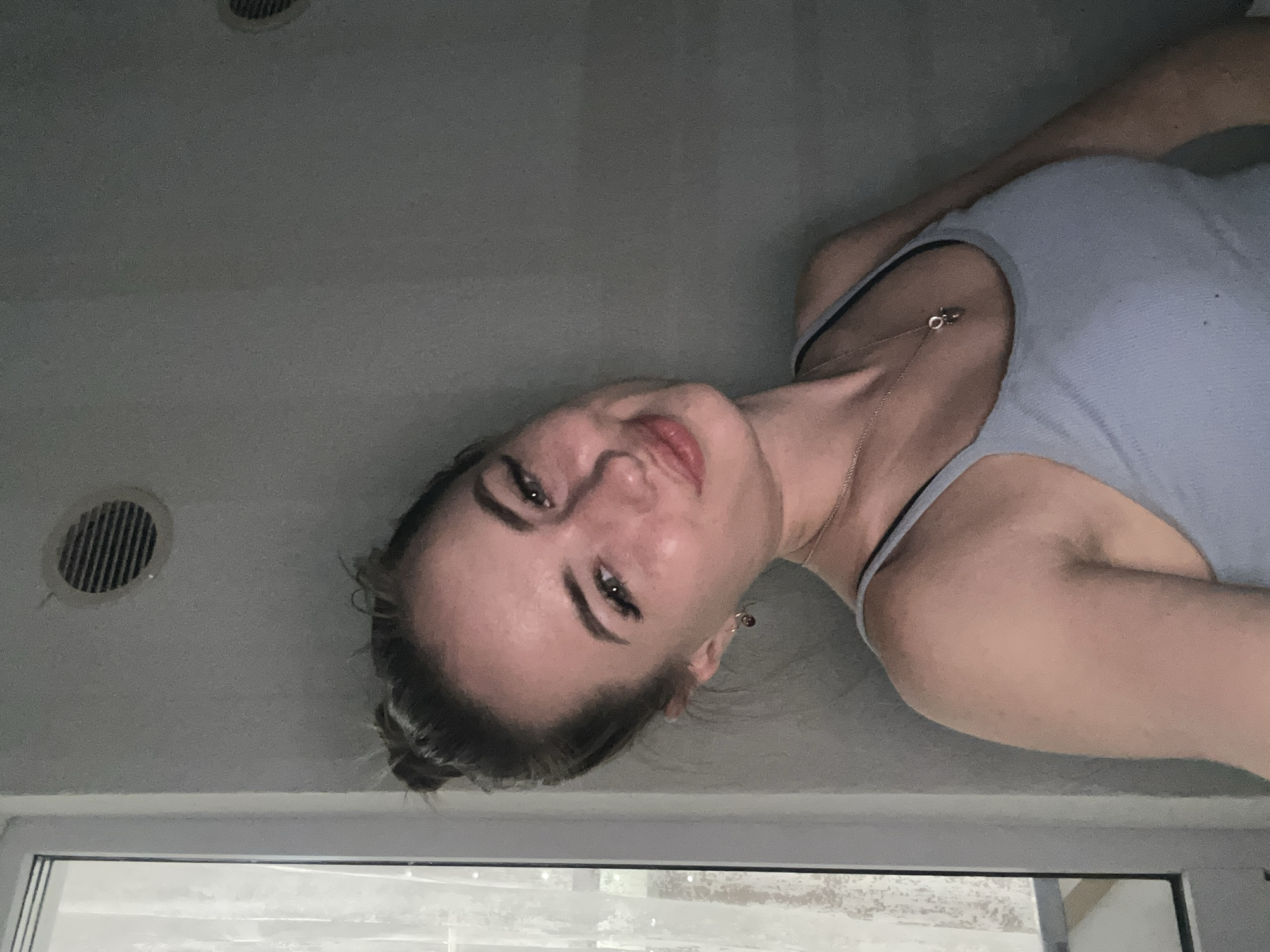 A young Caucasian woman with her hair pulled up in a bun is pictured from the chest up, looking directly at the camera with a slight smile. She is wearing a light blue tank top and a silver necklace with a pendant. The background is a plain gray wall with two circular vents visible. To the left of the woman is a partially visible window with sheer white curtains. The lighting is dim, casting shadows on the wall.