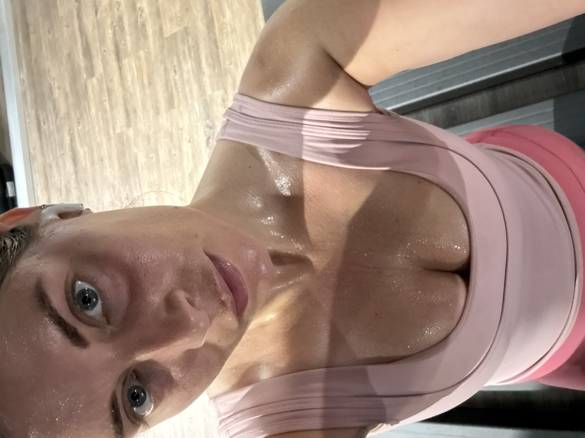 A close-up, high-angle selfie shows a woman wearing a pink tank top and matching leggings, likely after a workout. She has a white wireless earbud in her left ear. Her face and chest are covered in sweat. The background includes a wooden floor and a treadmill. There is no visible evidence to indicate that the image was taken in Paris, France.