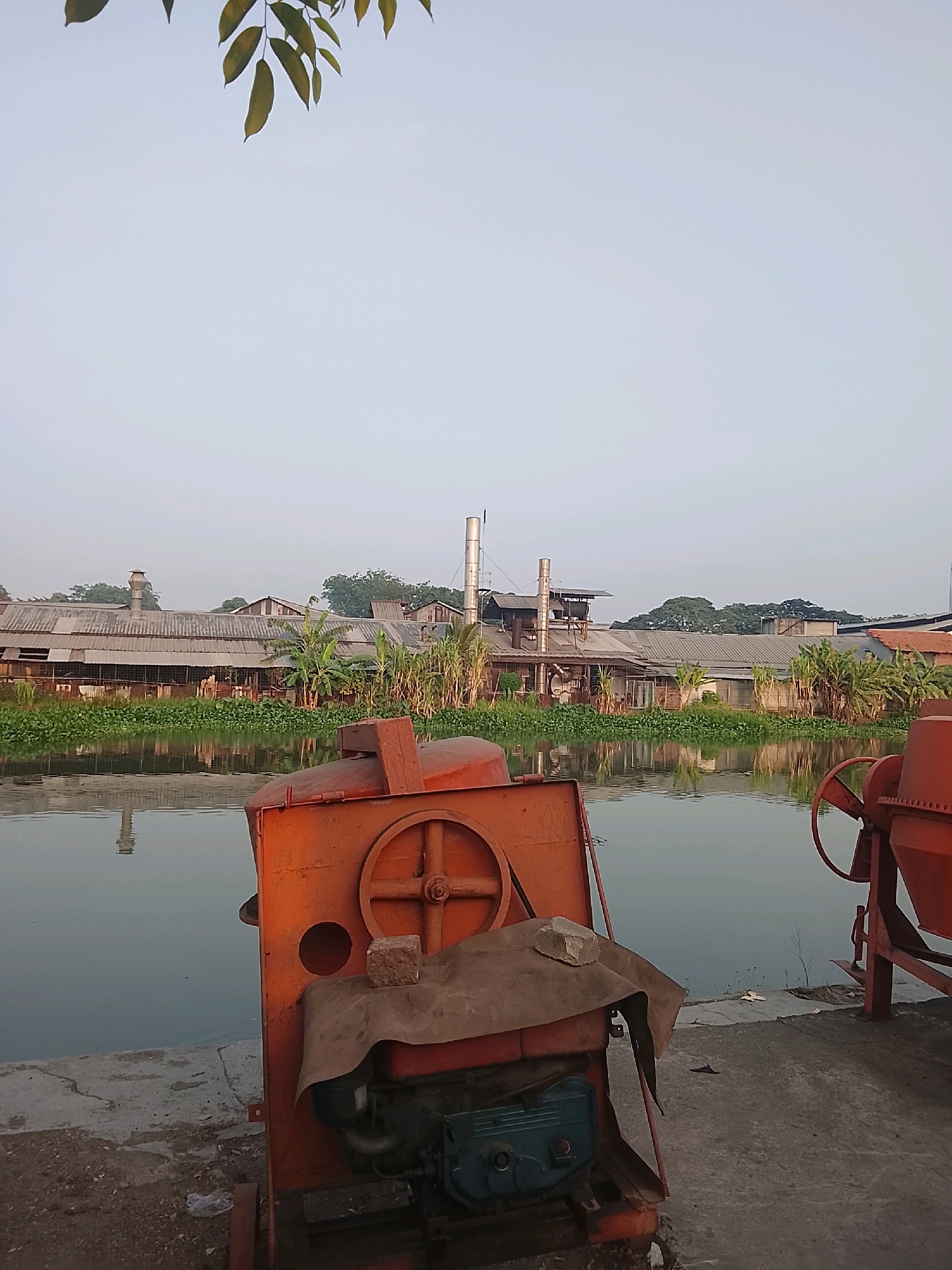 The image depicts an outdoor scene, likely in the industrial area of Surabaya, Indonesia. In the foreground, two orange cement mixers are visible. The one on the left is partially obscured by a tarp and has a small brick resting on it. The mixer on the right is seen from its side. A concrete walkway runs along the edge of a calm body of water. Across the water, a long, industrial building complex with a corrugated metal roof and several smokestacks stretches along the bank. Lush green vegetation, including what appear to be banana trees and reeds, grows between the water and the buildings. The sky is clear and pale, suggesting it might be early morning or late afternoon. The overall impression is one of industrial activity situated adjacent to a natural waterway.