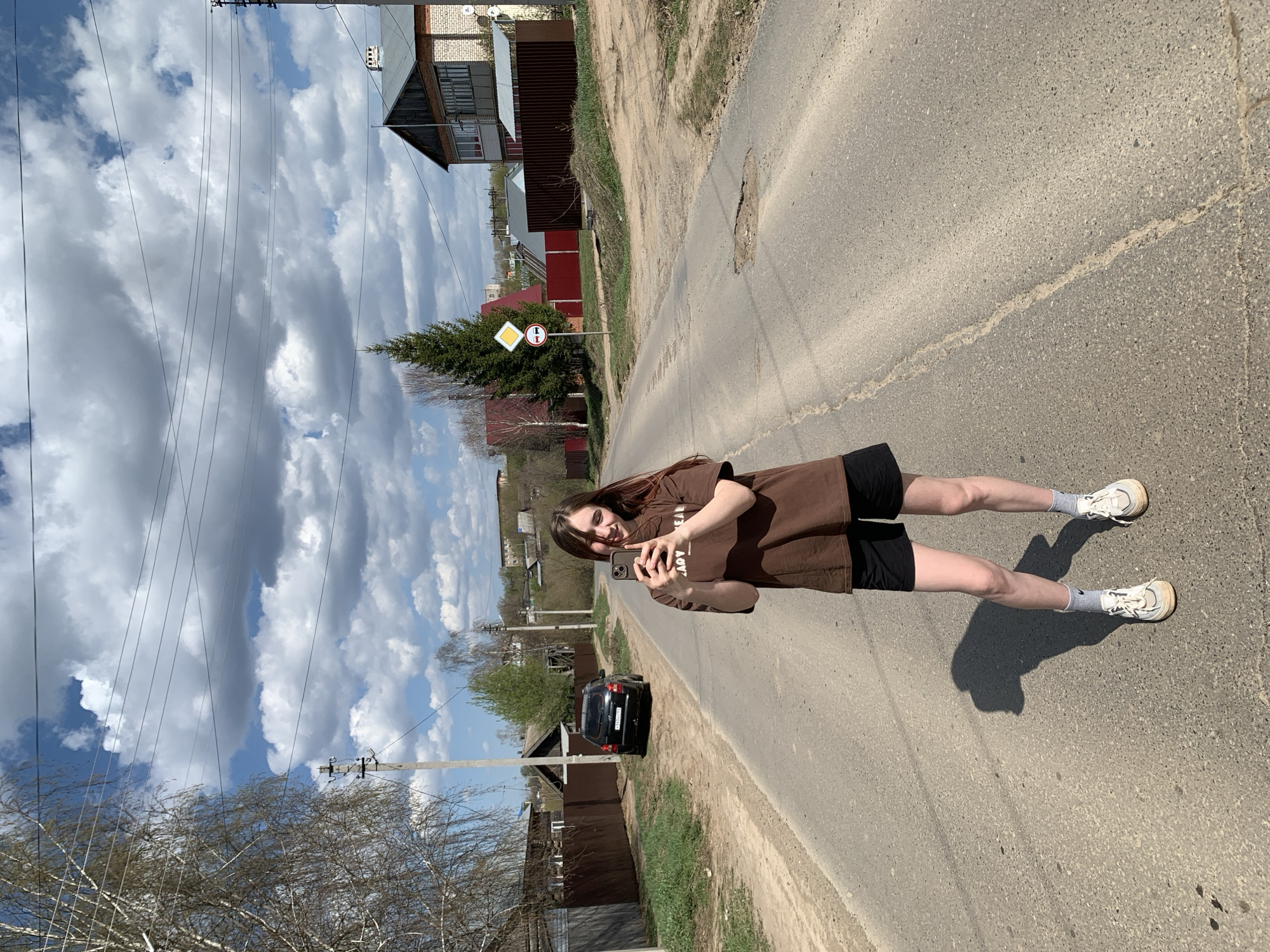 A young woman stands on an asphalt road holding a smartphone, seemingly taking a picture. She is wearing a brown t-shirt, black shorts, white socks, and white sneakers. The road is cracked and shows signs of wear. Houses and fences line the road, with trees scattered throughout the background. A black car is parked on the side of the road. The sky is partly cloudy. A road sign is visible indicating an intersection ahead. The location is identified as Фоки, Russia based on the presence of road signs and house styles typical of this region.