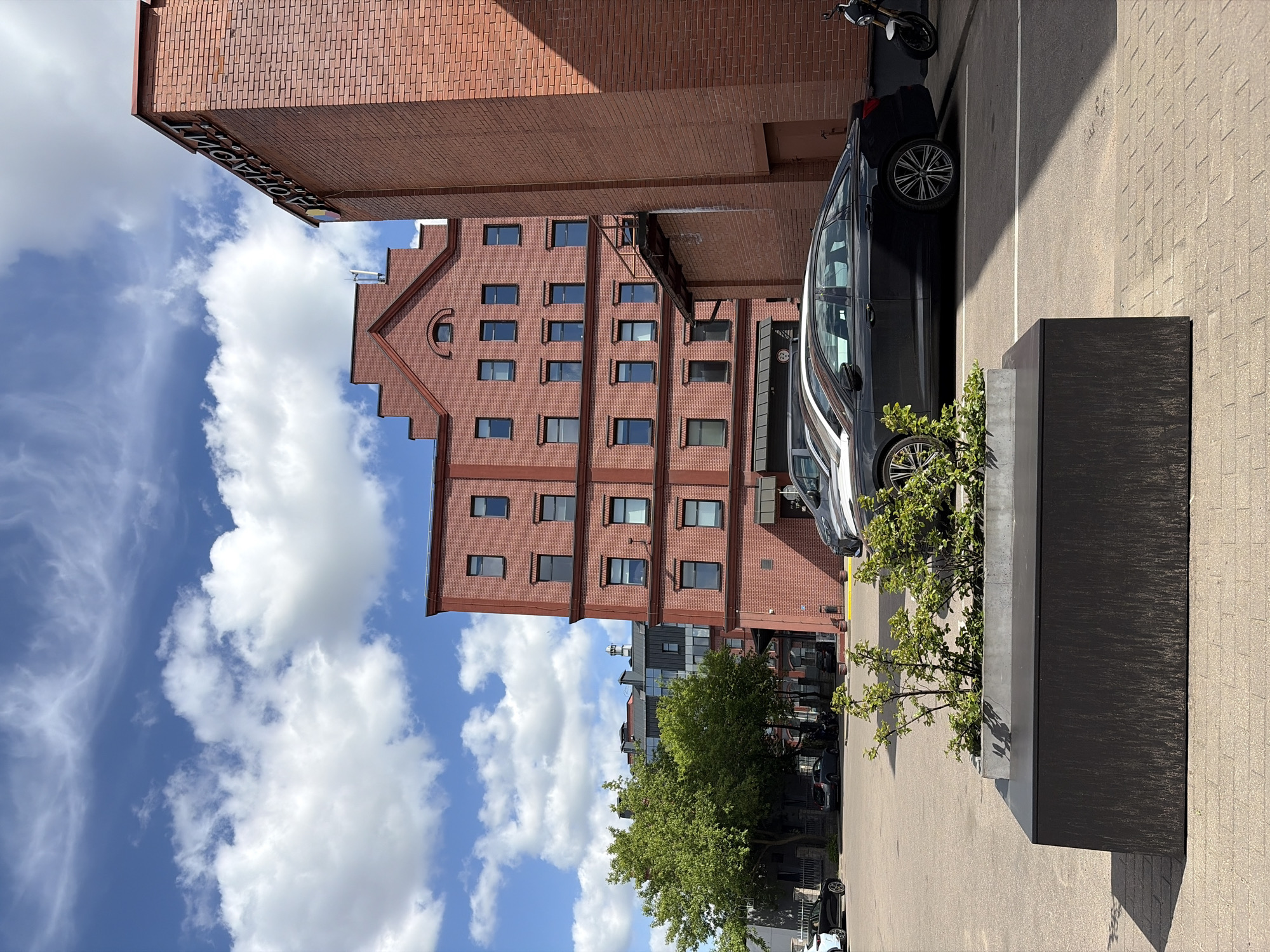 The image depicts an outdoor scene in Minsk, Belarus, featuring modern architecture. A multi-story red brick building with symmetrical windows and a distinctive stepped gable is the focal point. The building is located next to another brick building, also multi-storied, with similar architectural elements. A dark-colored car is parked adjacent to the brick buildings, partially obscured by a large, rectangular planter box filled with greenery. To the right of the car, the ground is paved with light-colored bricks. A glimpse of blue sky filled with clouds is visible in the background. A motorcycle is parked behind the car. Trees are also visible in the background, adding to the urban landscape.