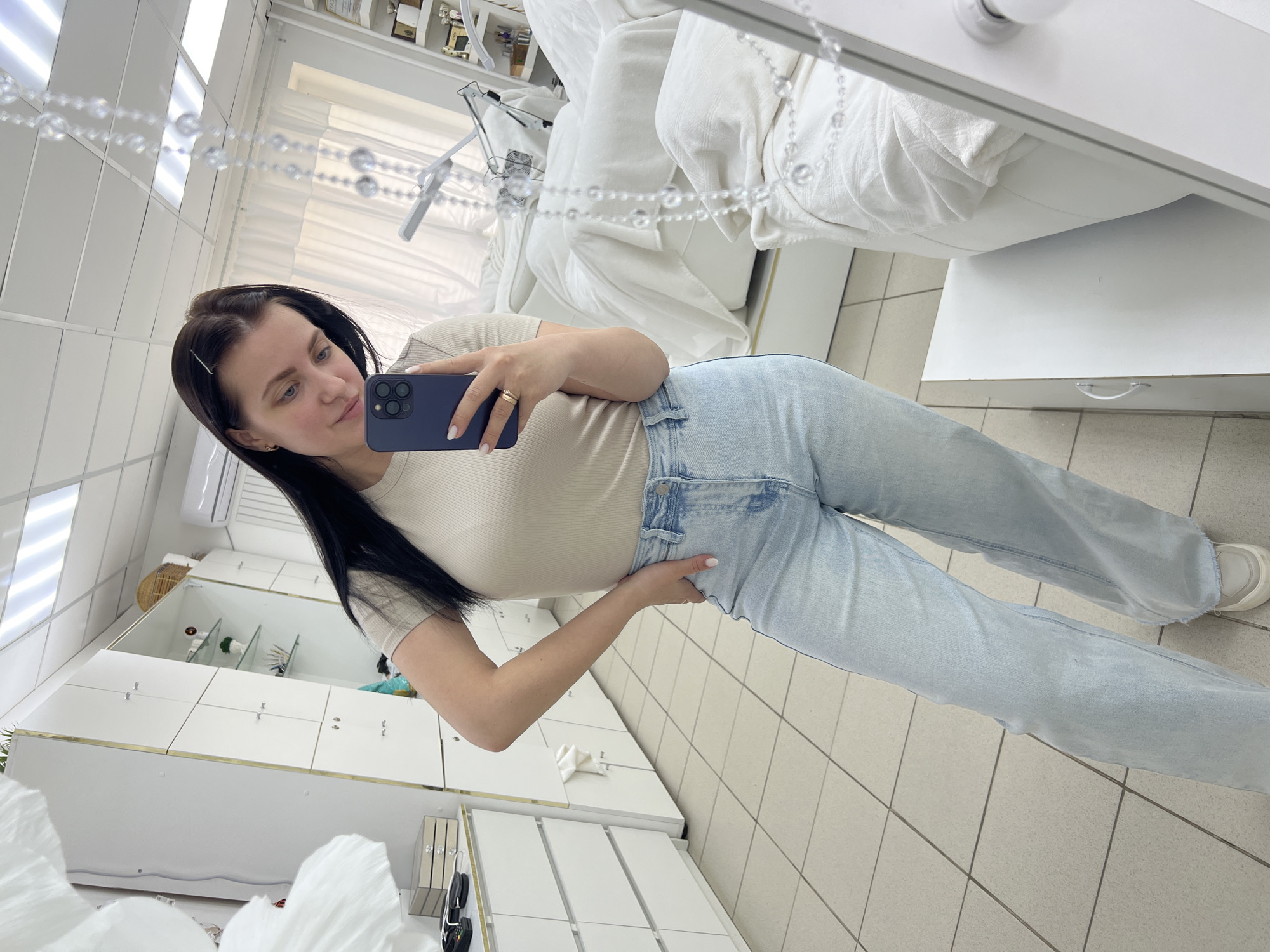 A woman is taking a selfie in a room, likely a salon, in Chaikovsky, Russia. She is holding a blue smartphone and is wearing a beige fitted shirt and light blue jeans. The room has white cabinets and a white examination table with a white cover. The floor is tiled in a light color.