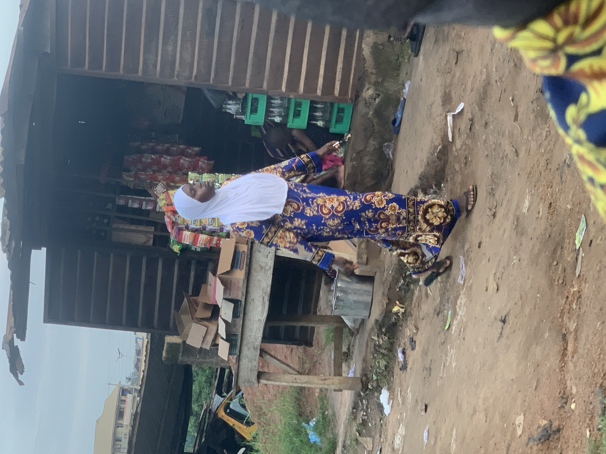 A woman wearing a white hijab and a blue and gold floral patterned dress walks on a dirt path in Shagamu, Nigeria. She is walking past a makeshift wooden structure that appears to be a small shop. The shop is open and contains shelves stocked with bottles, some in green crates. Bags of snacks are hanging from the front of the shop. In front of the shop, there is a wooden table with cardboard boxes on it. The ground is covered in dirt and debris, including plastic bags and wrappers. In the background, a yellow vehicle is partially visible. The sky is overcast. The scene appears to be a street market or a roadside vendor area.