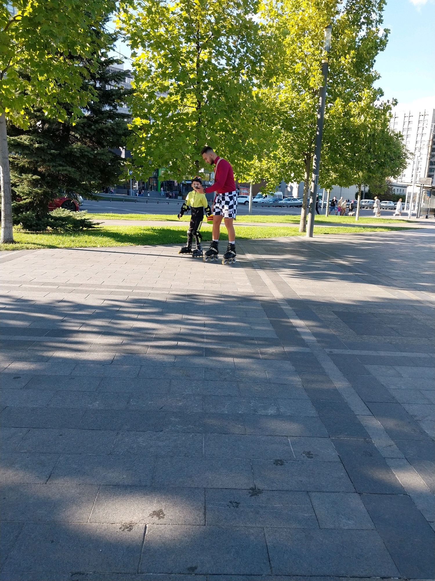 The image depicts an outdoor scene in the city of Dnipro, Ukraine. Two individuals, an adult and a child, are rollerblading on a paved pathway. The adult, wearing a red long-sleeved shirt and black and white checkered shorts, stands on the right. The child, dressed in a yellow and black jacket and pants, is positioned to the left and slightly behind the adult. Both are wearing rollerblades. The pathway is paved with large gray tiles, and shadows from nearby trees are cast across it. Behind the pathway, a strip of green grass is visible, beyond which is a road with traffic. Trees with lush green foliage line the area, and modern buildings can be seen in the background. The sky is blue with some scattered clouds, indicating a clear day.