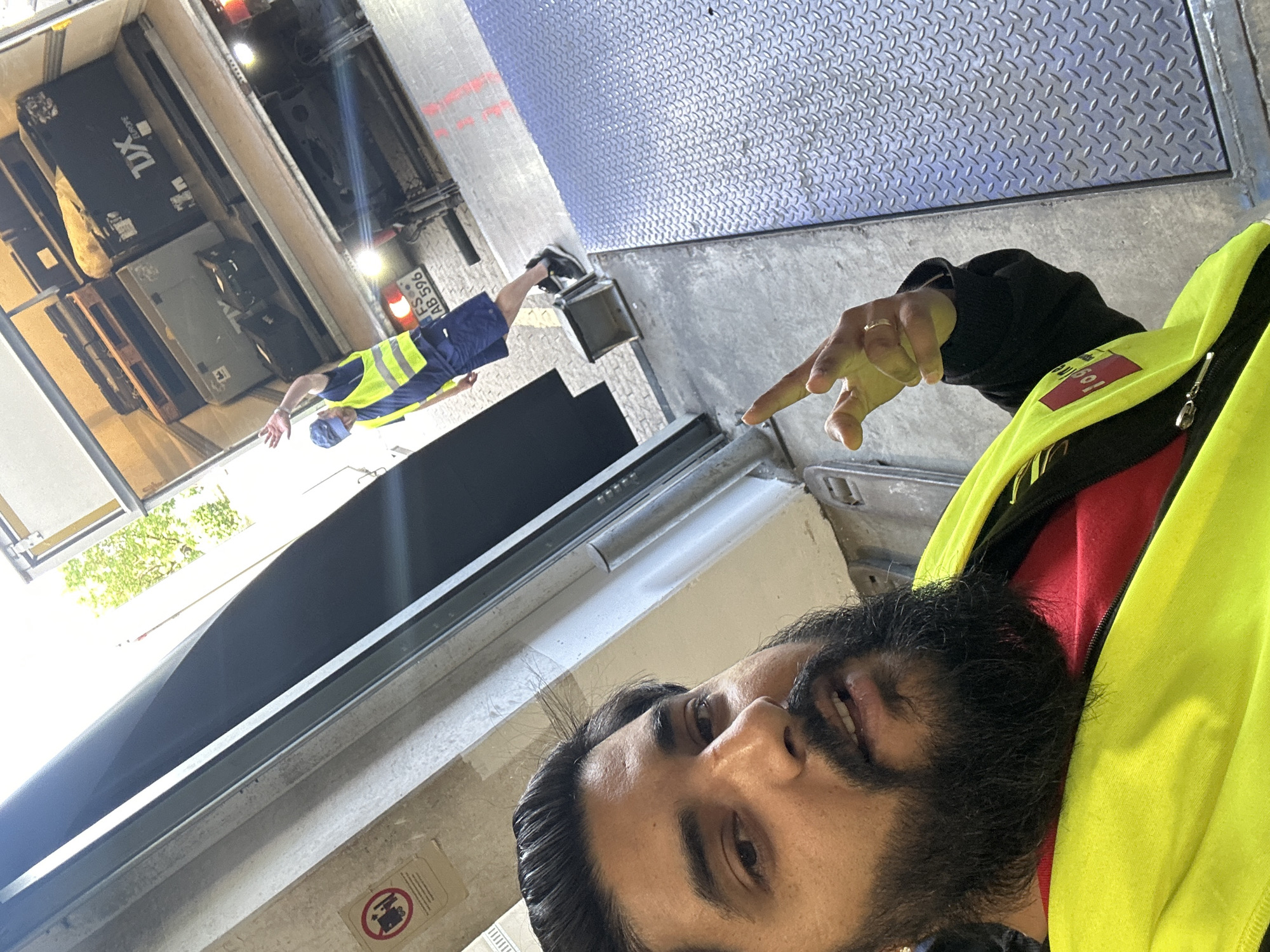 A person in a bright yellow safety vest and red shirt is posing for a selfie in a loading dock area. The person has dark hair and a beard and is making a peace sign with their right hand. Behind them, a truck is partially visible with its rear doors open, loaded with various boxes and pallets. Another person wearing a blue cap and a yellow safety vest is standing in the back of the truck, waving. The location appears to be an industrial or logistics facility, possibly in Schweitenkirchen, Germany, given the license plate on the truck which reads "FS AB 596". The ground is concrete and metal, with some red markings visible. A "no forklifts" sign is mounted on a wall to the left. The overall lighting suggests it is daytime, with sunlight filtering in.
