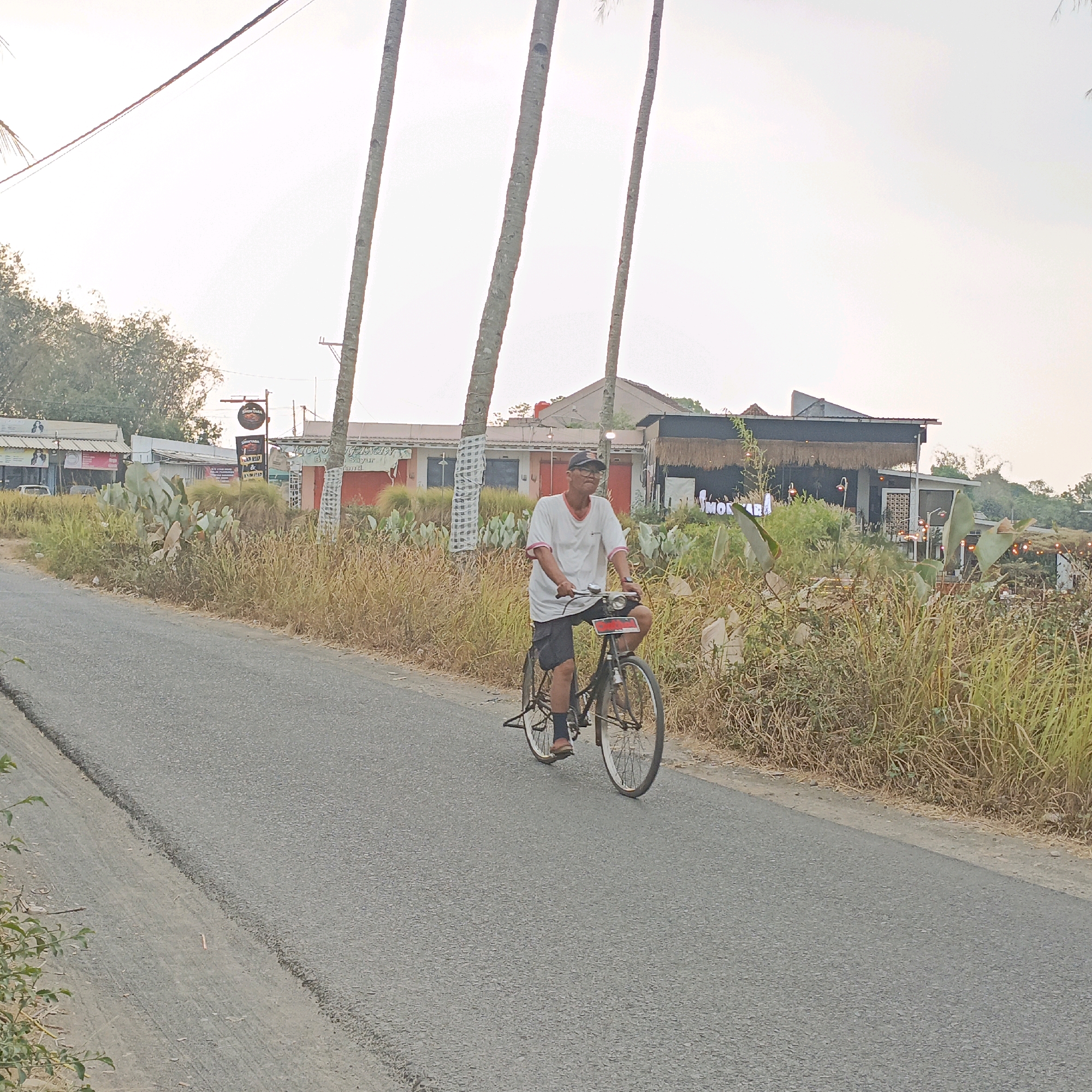 In Jombor, Indonesia, a man rides a bicycle on a paved road. The man is wearing a white t-shirt with pink trim, dark shorts, and a baseball cap. He is looking forward as he cycles. The road is flanked by dry grass and vegetation. In the background, there are several buildings with thatched roofs, some trees, and utility poles. The sky is bright and hazy.